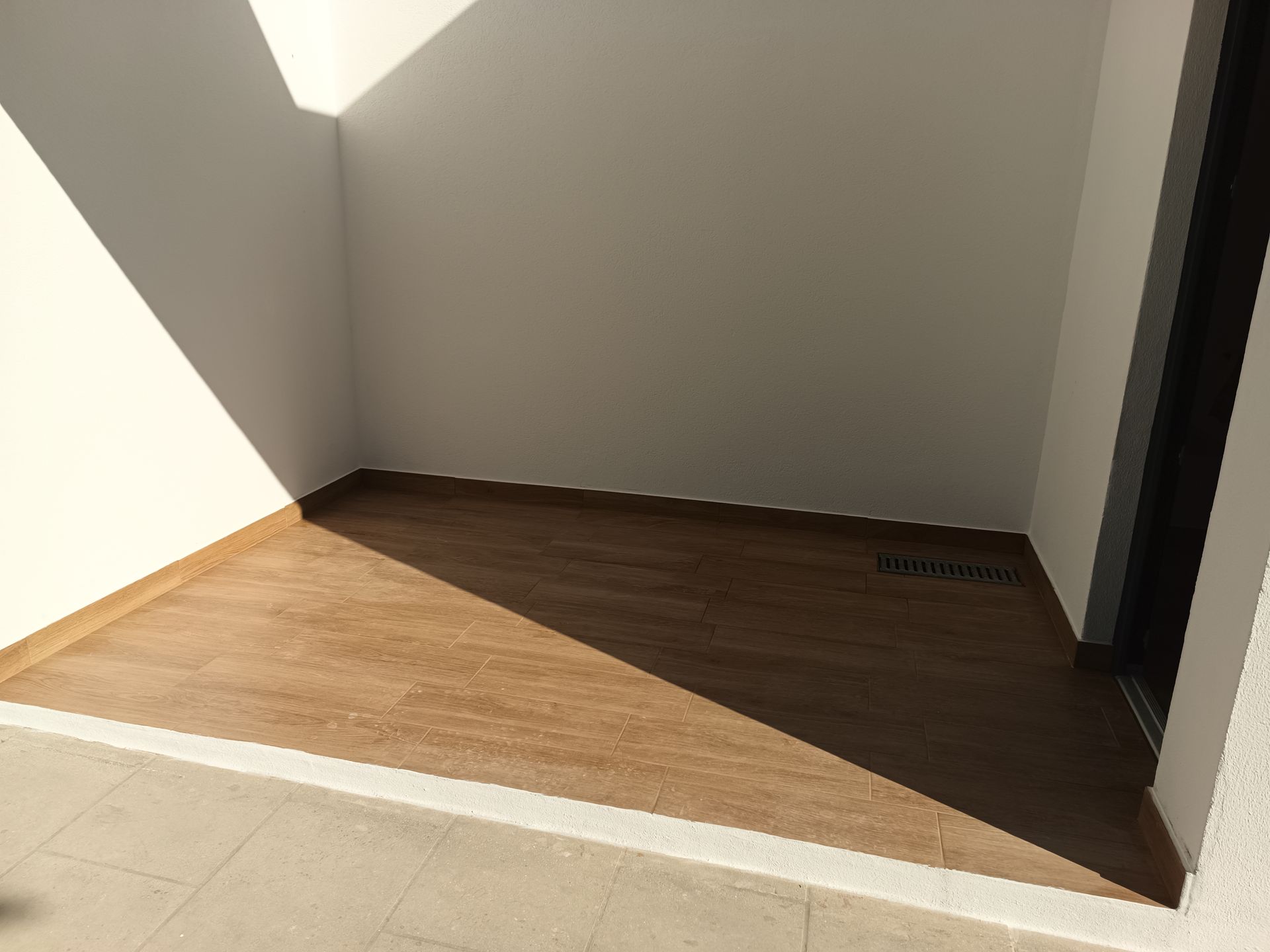 Uma pequena varanda vazia com piso que imita madeira e paredes brancas, parcialmente iluminada pela luz do sol.