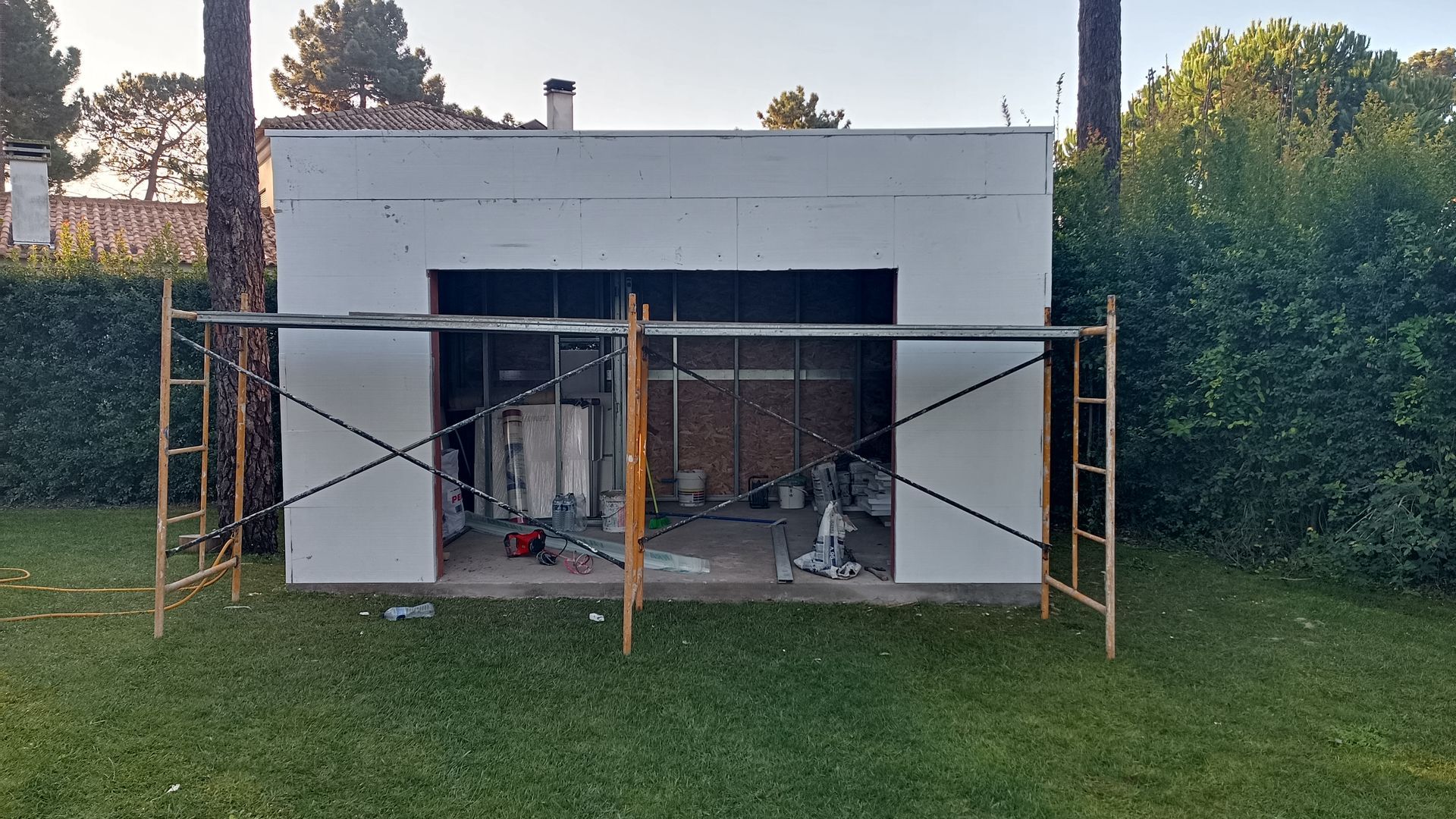 Prédio em construção com andaimes em um gramado. Exterior branco, porta aberta, ferramentas visíveis no interior.