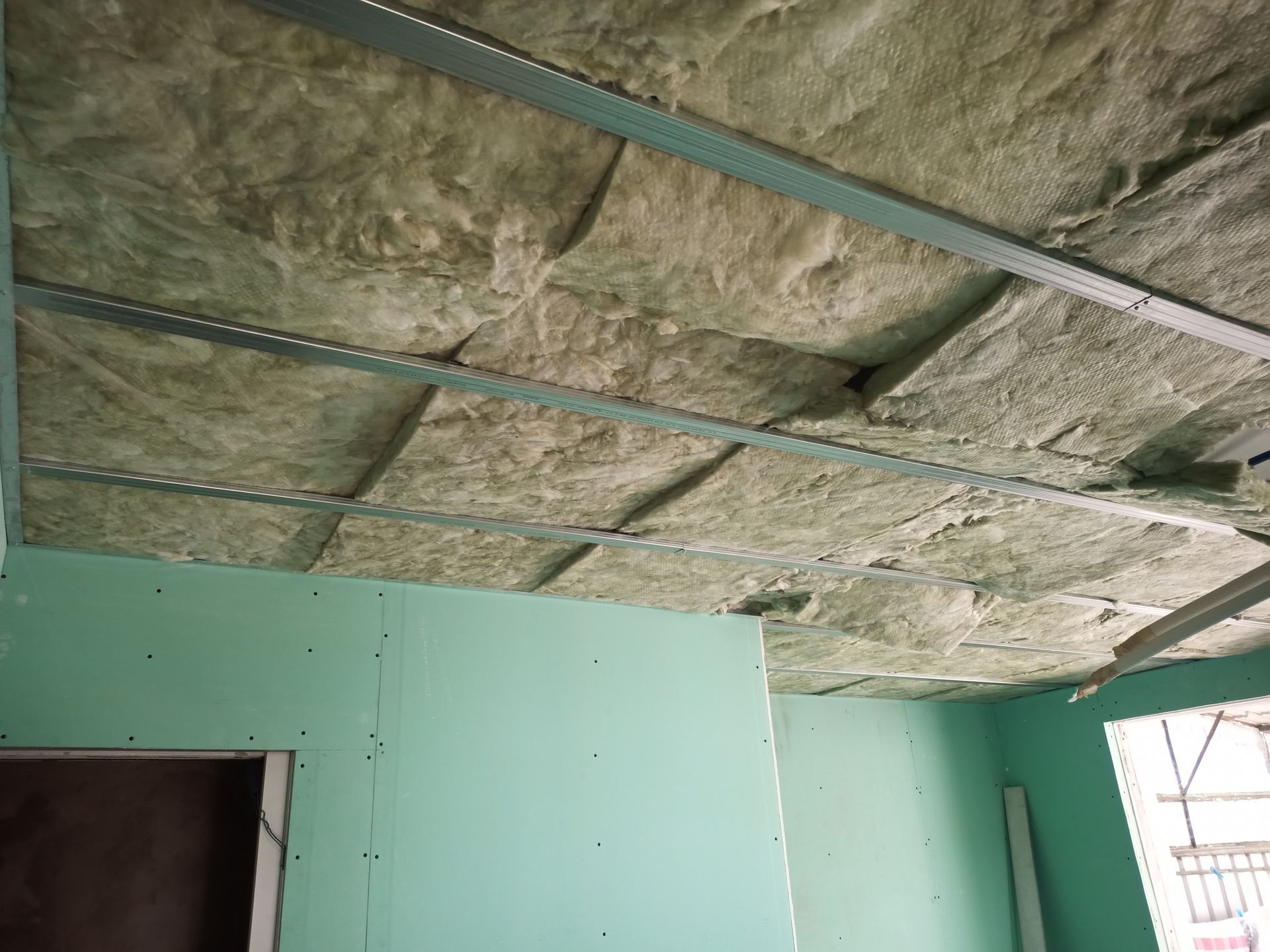 Teto com isolamento entre suportes metálicos; paredes de gesso cartonado. Tons de verde e marrom. Cena de construção.