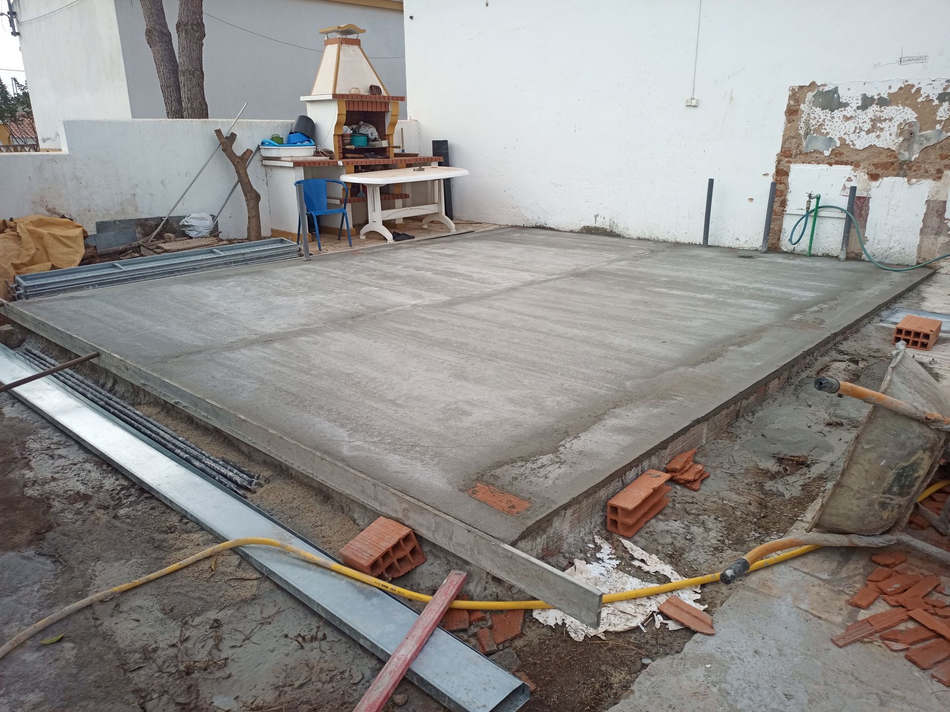 Pátio de concreto recém-construído, cercado por tijolos e materiais de construção, próximo a uma parede branca e cozinha externa.