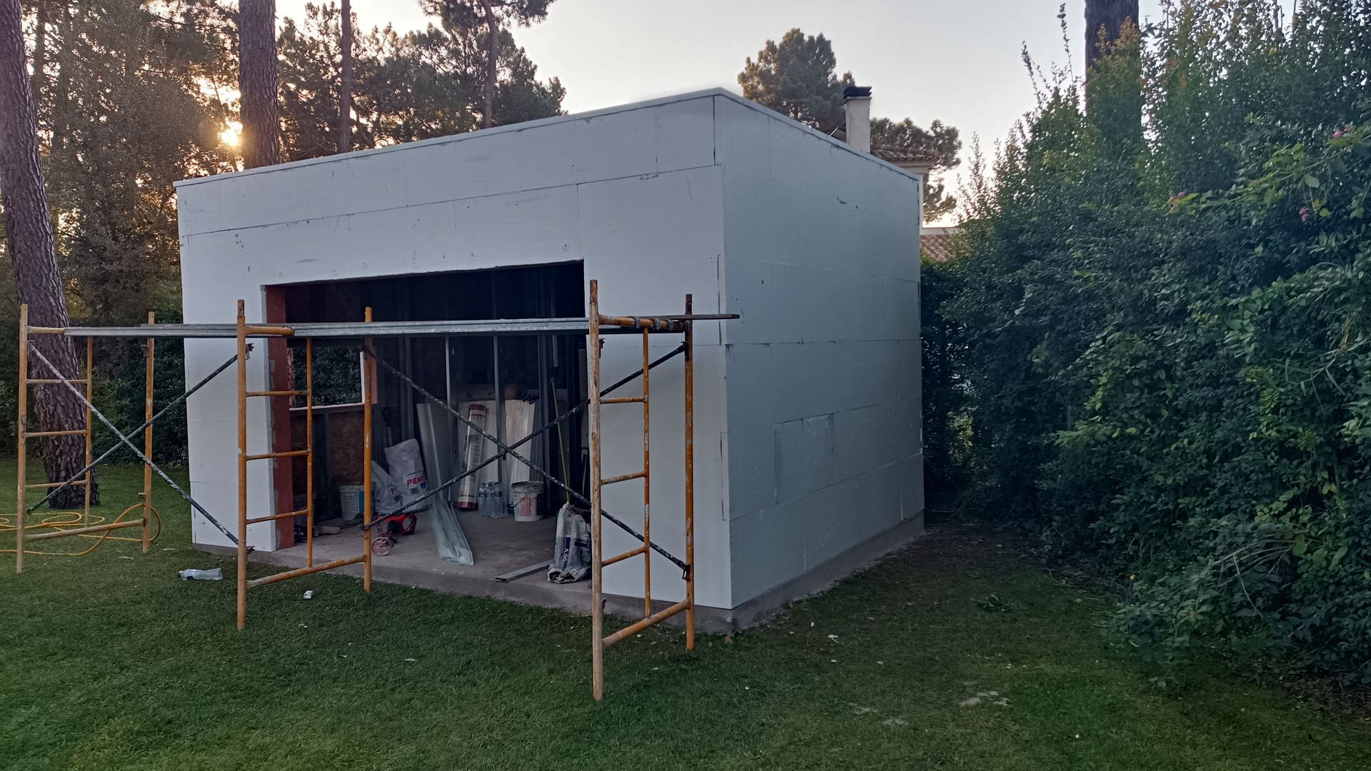 Prédio branco e quadrado em construção, com andaimes na frente. Grama verde e árvores ao fundo.