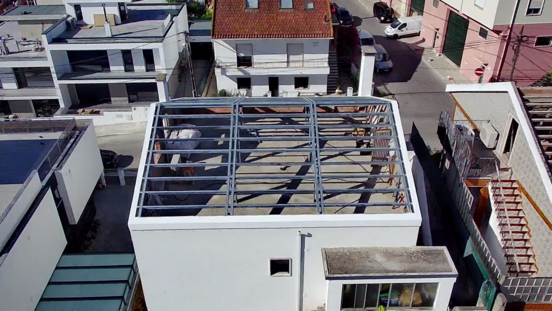 Vista aérea de um edifício branco com estrutura metálica do telhado exposta, outros edifícios e a rua ao fundo.
