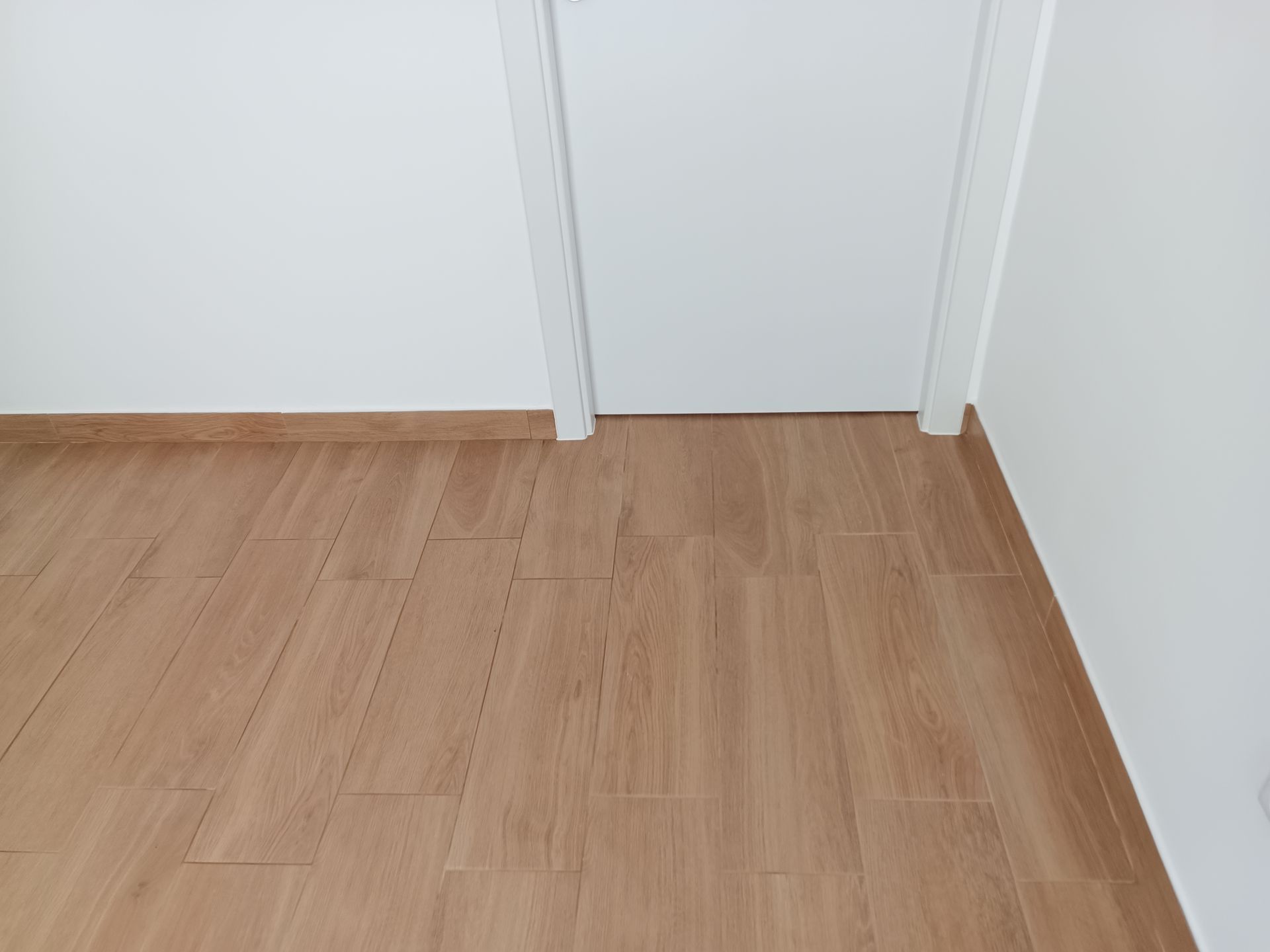 Quarto de canto com piso que imita madeira clara, paredes brancas e porta branca.