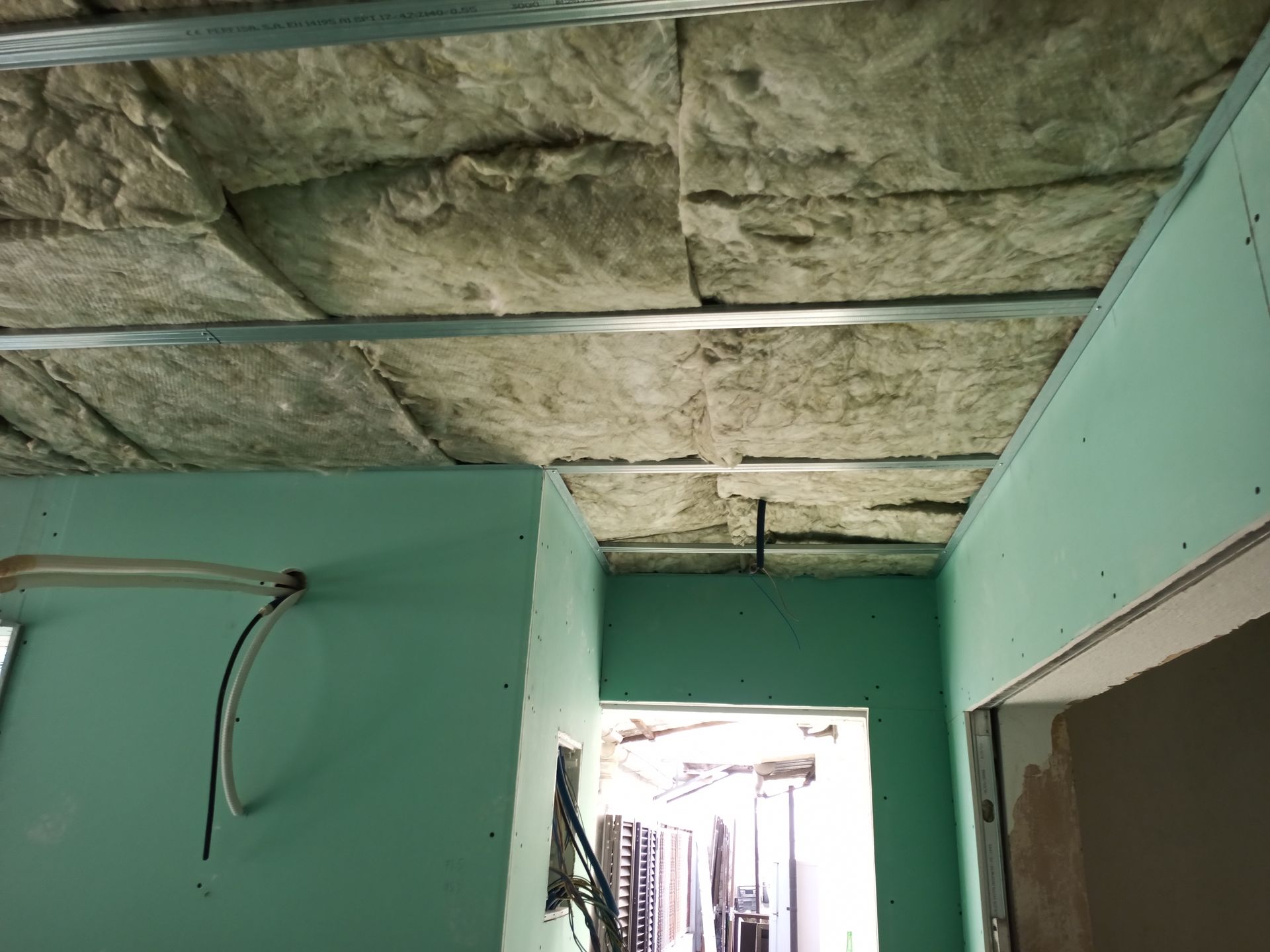 Isolamento de teto instalado entre suportes metálicos, adjacente à placa de gesso verde.