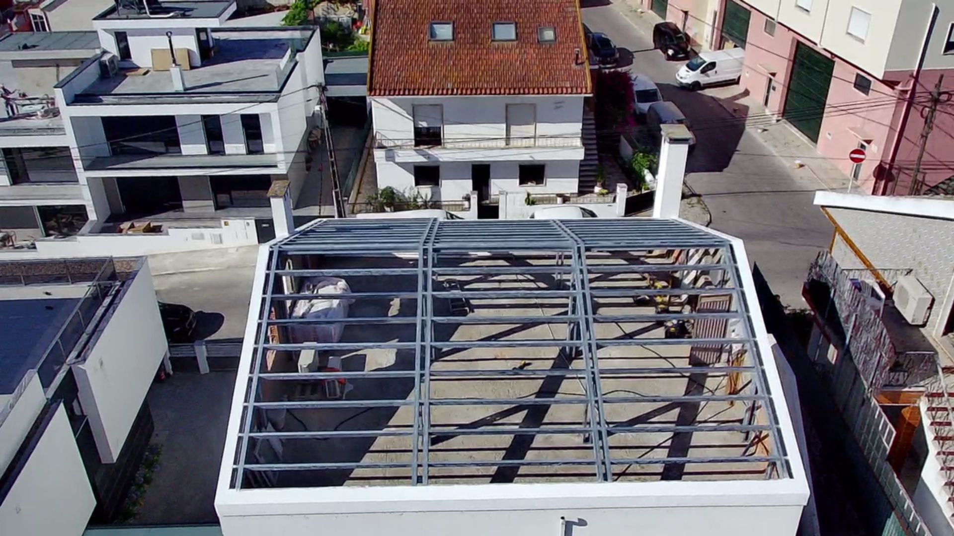 Vista aérea da cobertura de um edifício em construção com estrutura metálica, cercada por outros edifícios e uma rua.