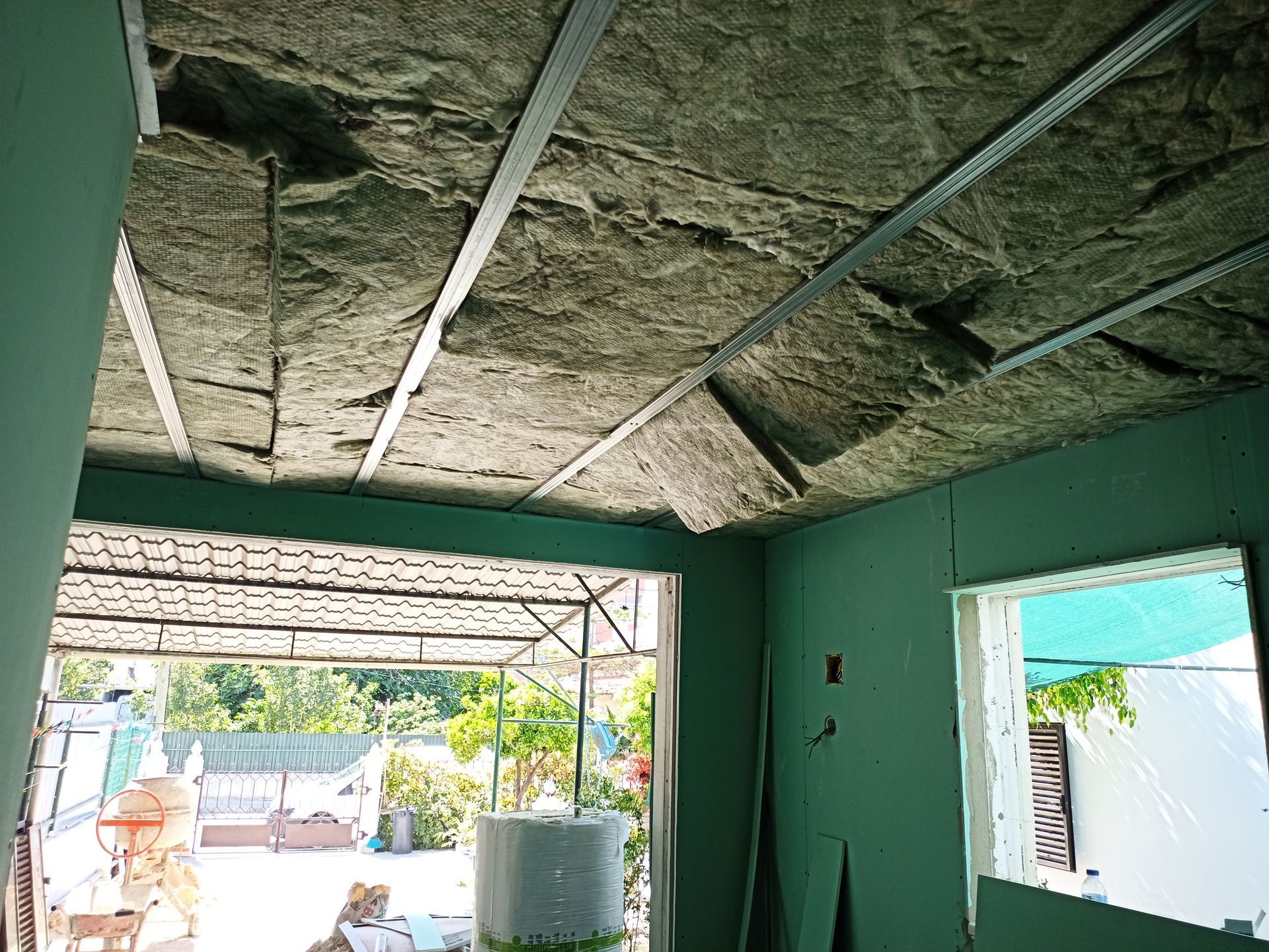 Vista interna de uma sala em construção com isolamento entre as vigas metálicas do teto, paredes verdes e uma porta aberta.