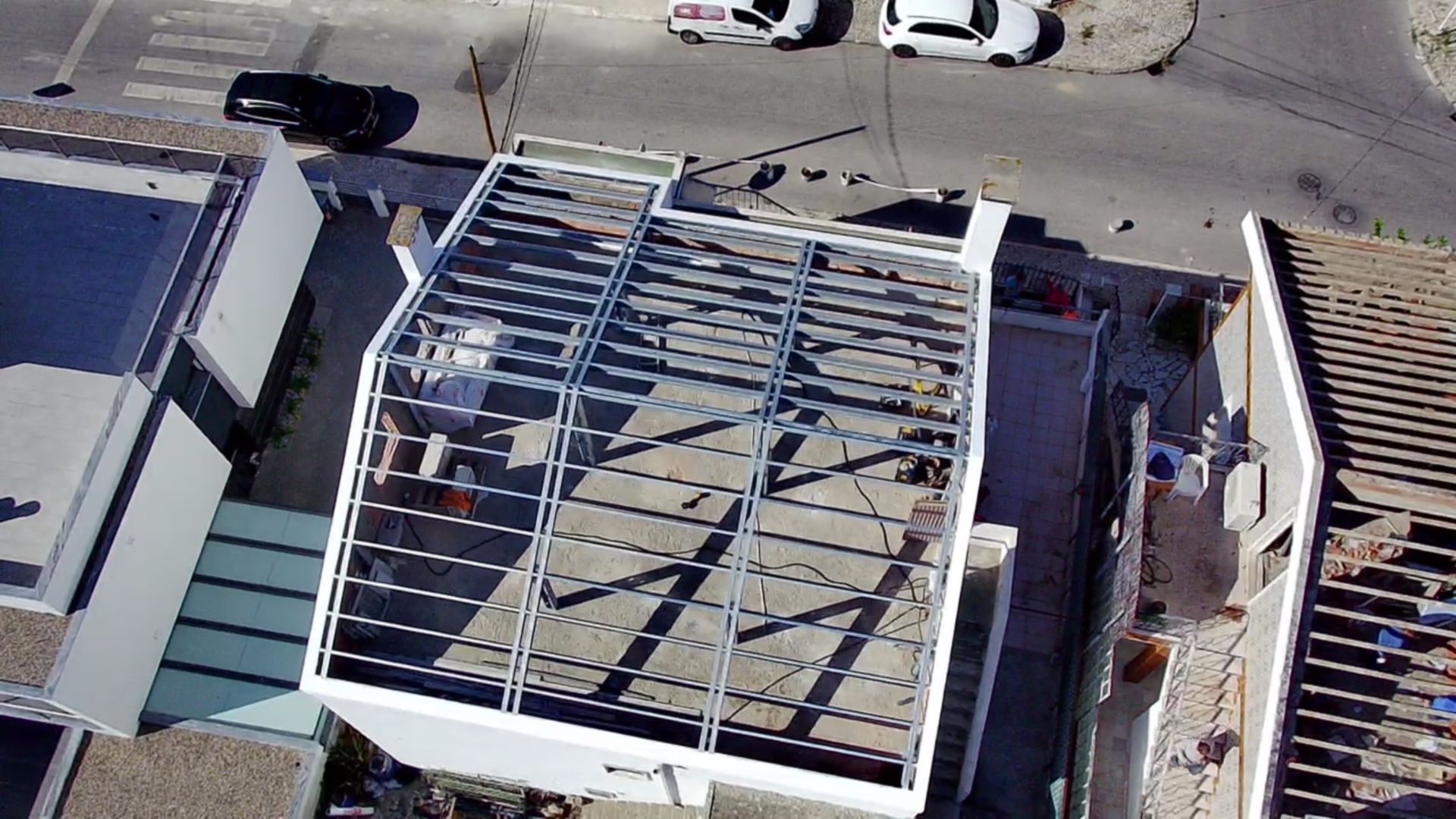 Vista aérea de um edifício em construção, com cobertura de estrutura metálica, sombras, rua e carros ao fundo.