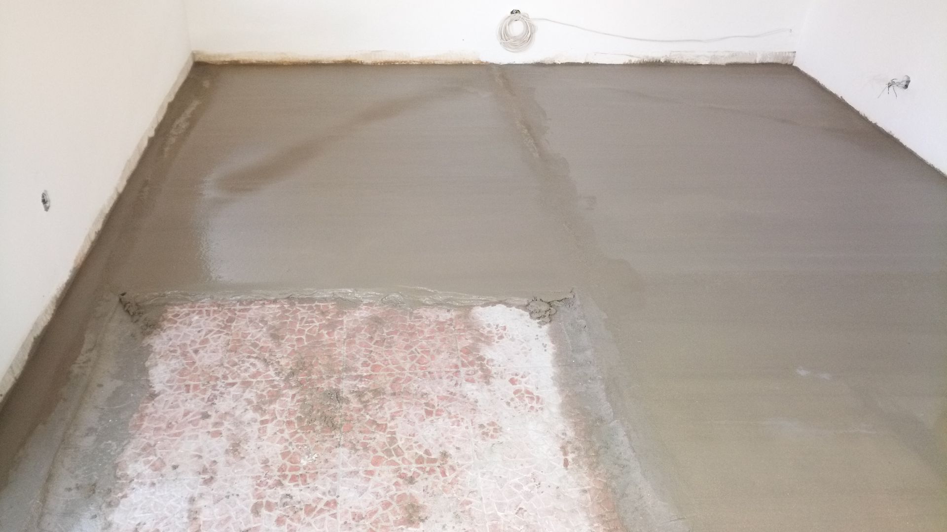 Piso de concreto cinza recém-moldado em um cômodo, cobrindo seções do piso antigo, com as paredes visíveis.