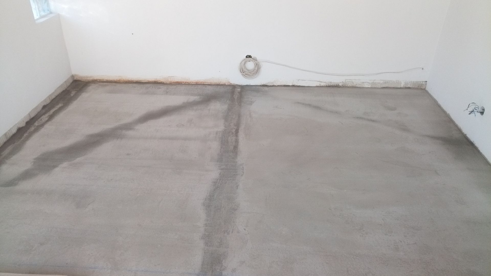 Piso de concreto em um cômodo com paredes brancas, inacabado, com luz vinda de uma janela.
