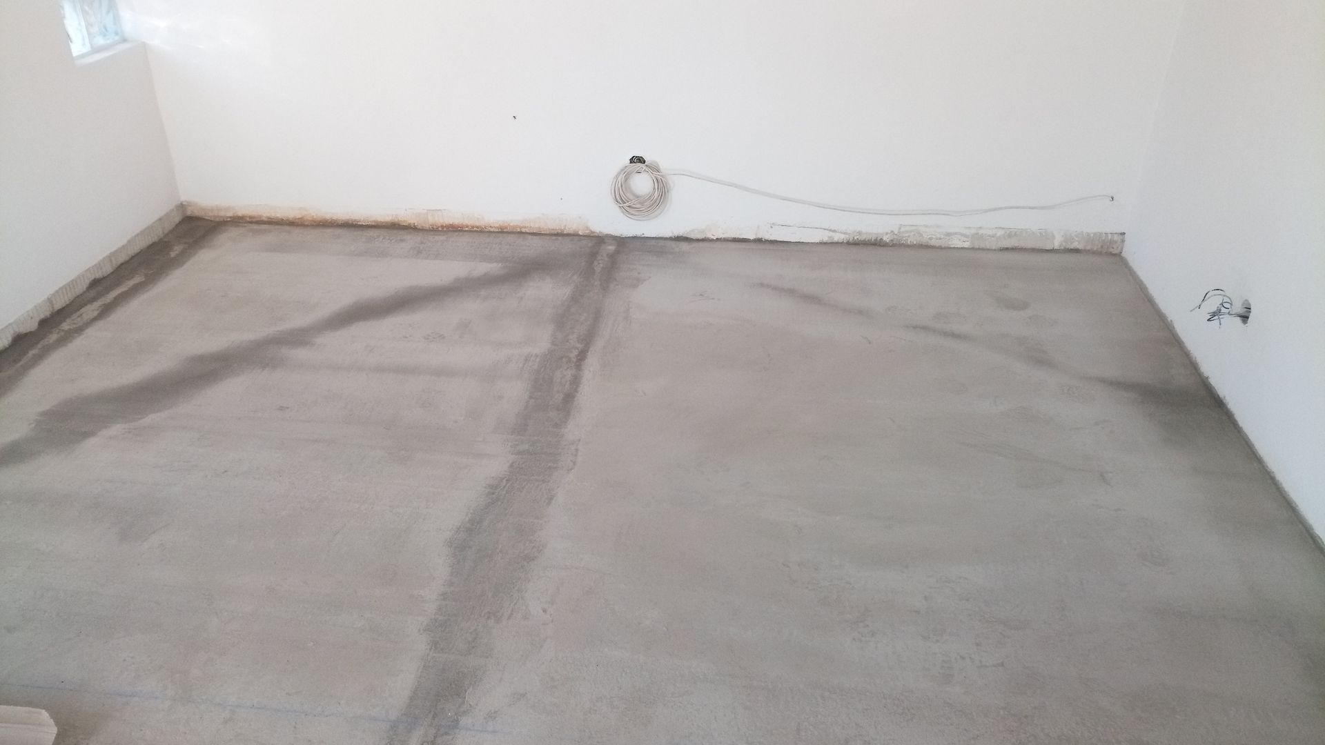 Piso de concreto cinza em um cômodo com paredes brancas, sem acabamento. Fiação elétrica na parede.