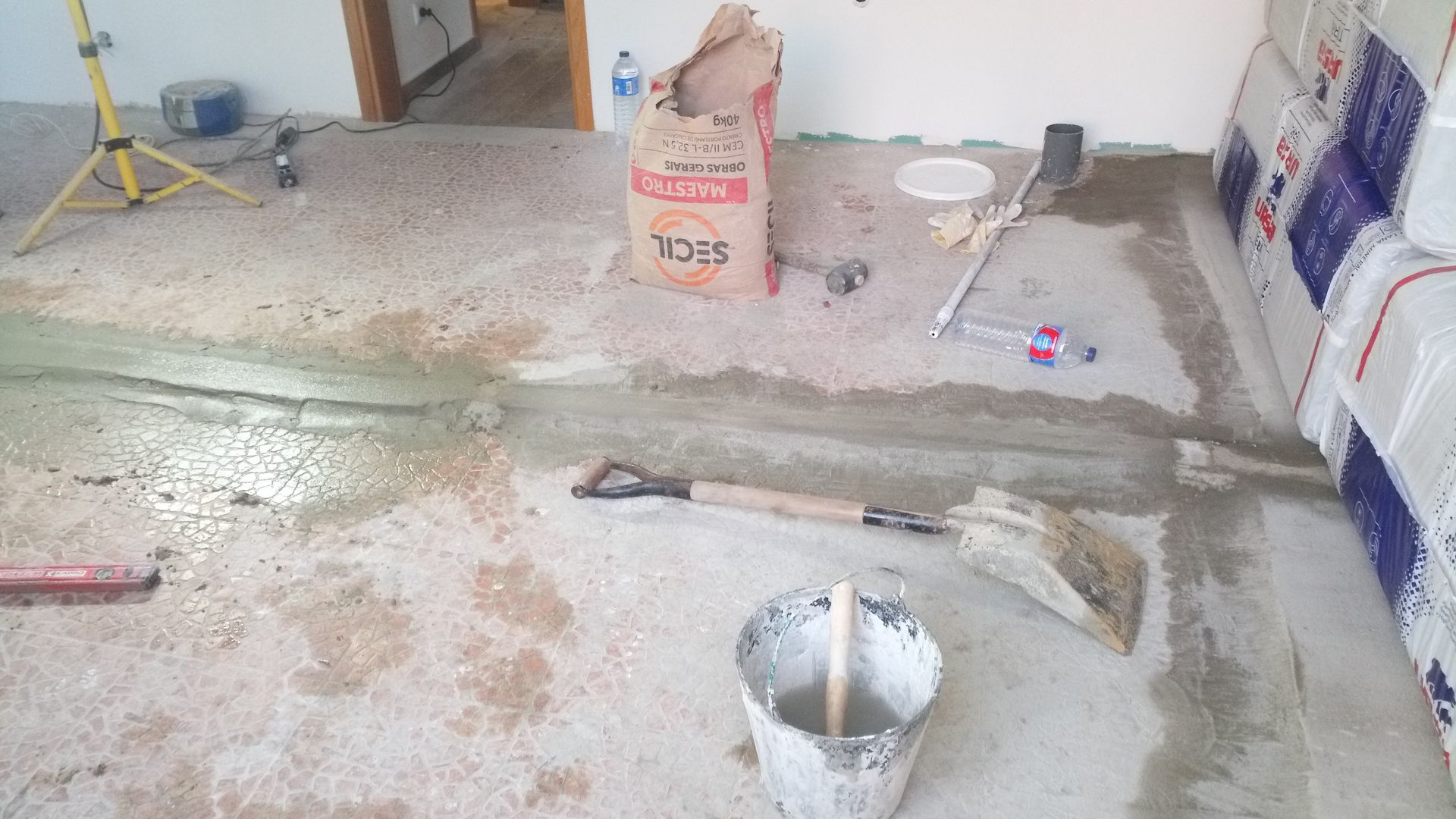 Canteiro de obras: Saco de cimento, pá, balde e reparo do piso em andamento.