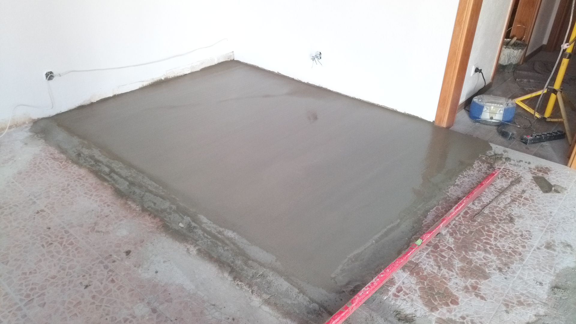 Seção de piso de concreto cinza recém-moldado. Cimento nivelado, próximo à porta e à parede.