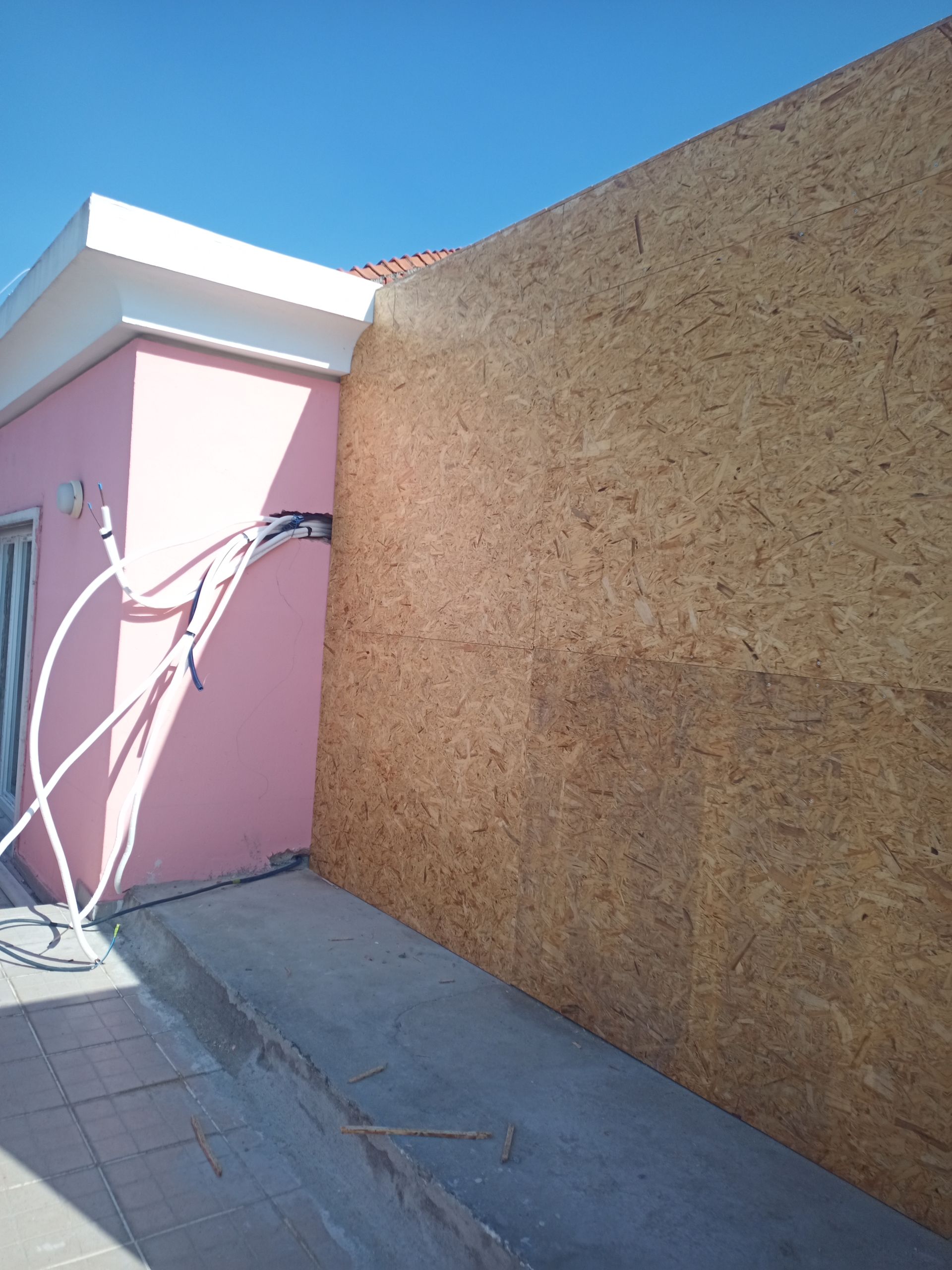 Um prédio rosa com detalhes brancos, parede bege e tubulações de serviços públicos contra um céu azul.