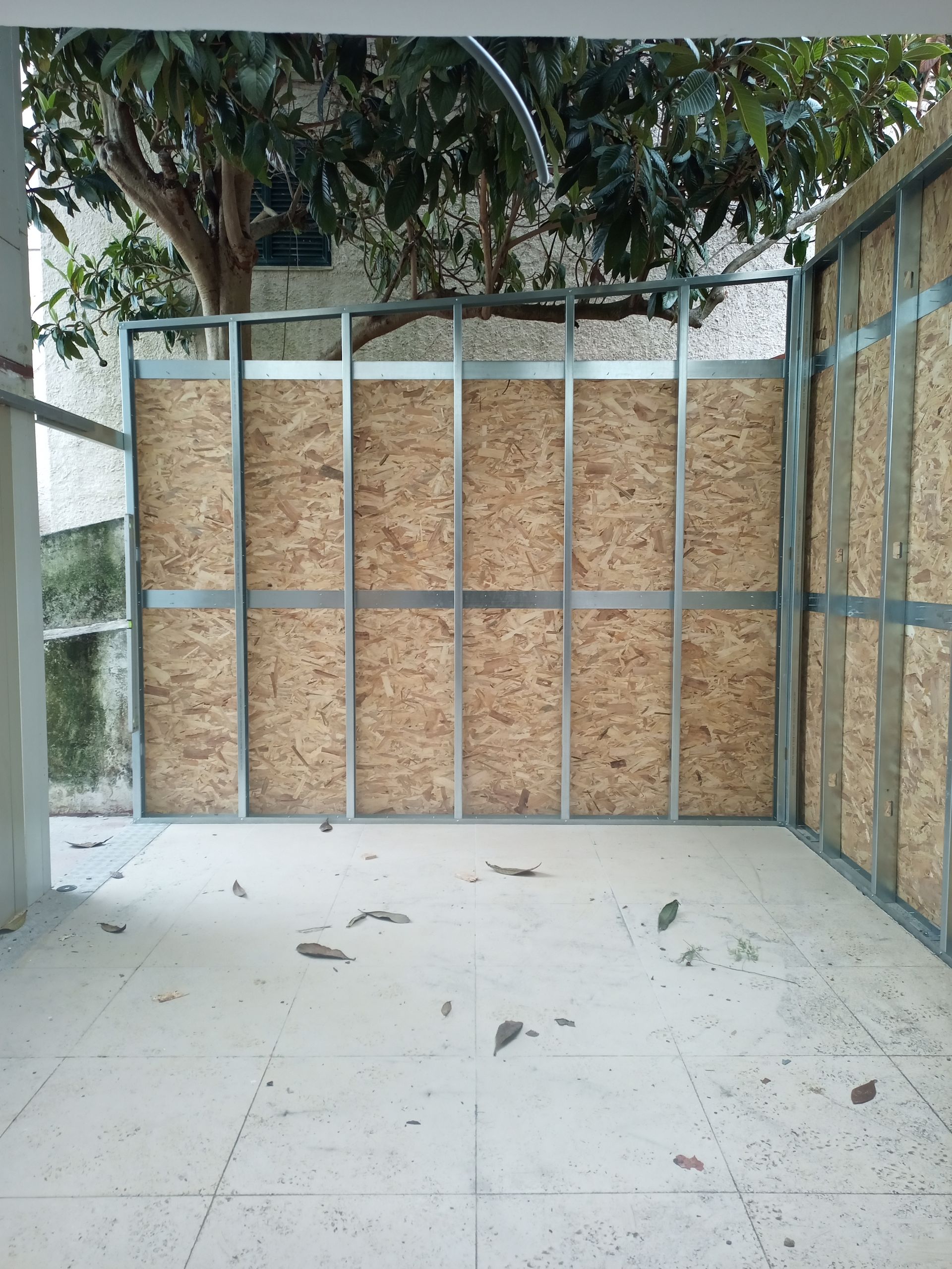 Canteiro de obras: estrutura metálica e placas de OSB formando uma parede parcial, entulho no piso de concreto, árvore visível do lado de fora.