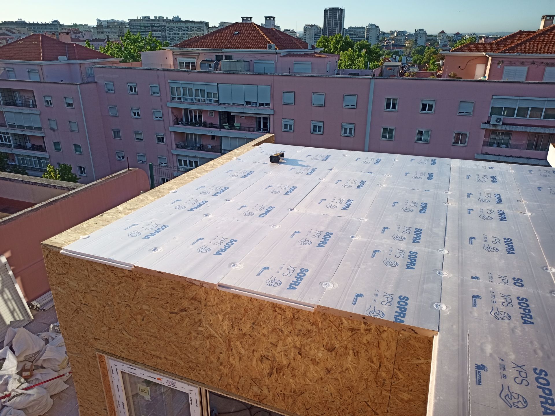 Telhado de placas OSB sendo coberto com material de cobertura Supra, com prédios cor-de-rosa ao fundo.