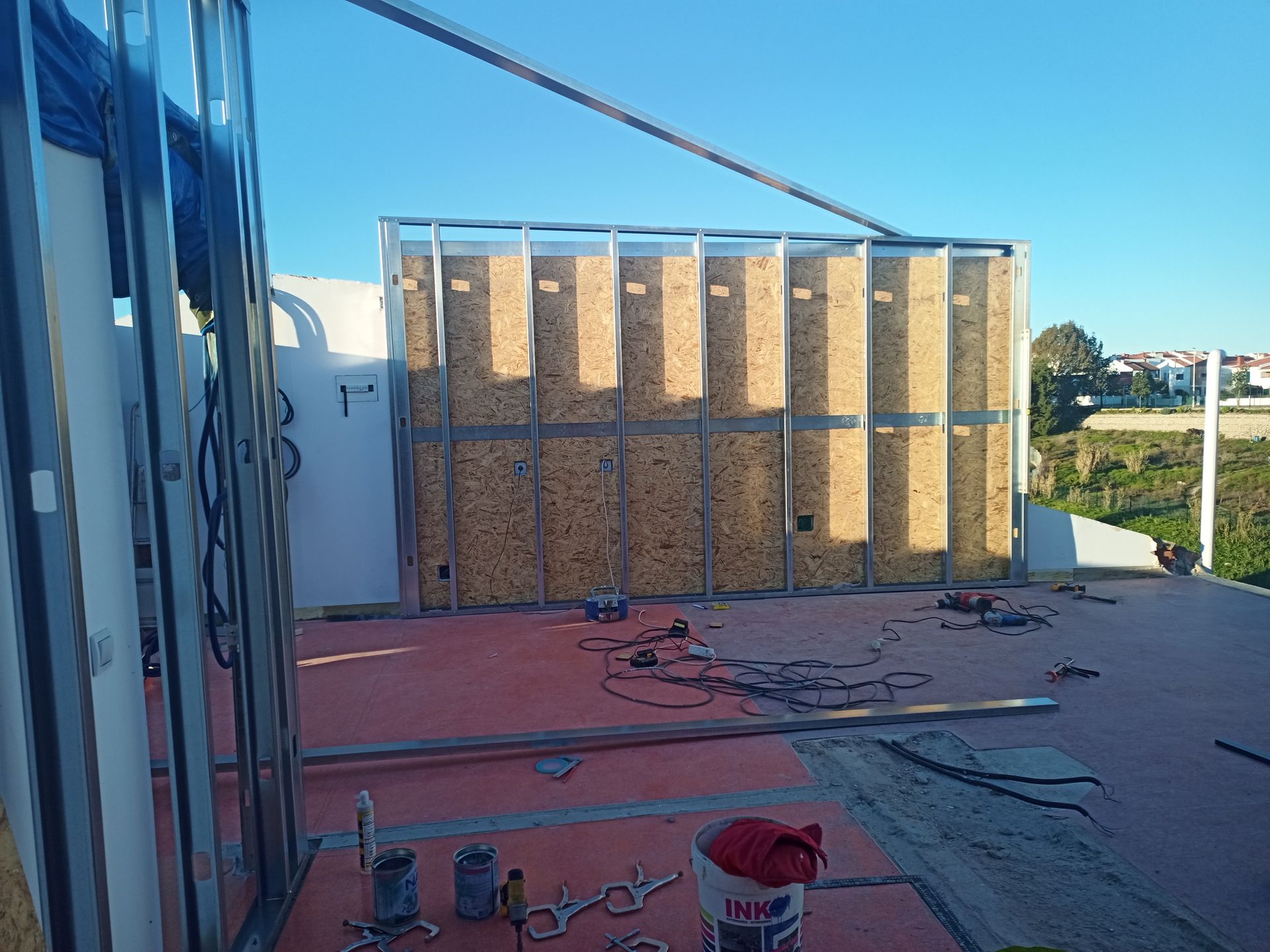 Canteiro de obras com uma parede de estrutura metálica preenchida com isolamento de madeira sobre uma superfície vermelha sob um céu azul.