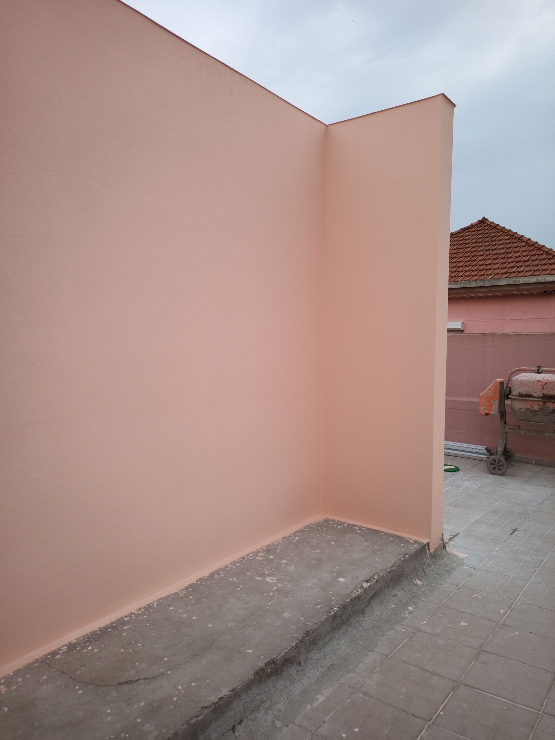 Paredes cor de pêssego se encontram em um ângulo reto, com uma saliência de concreto em primeiro plano. Um prédio com telhado de telha vermelha é visível.