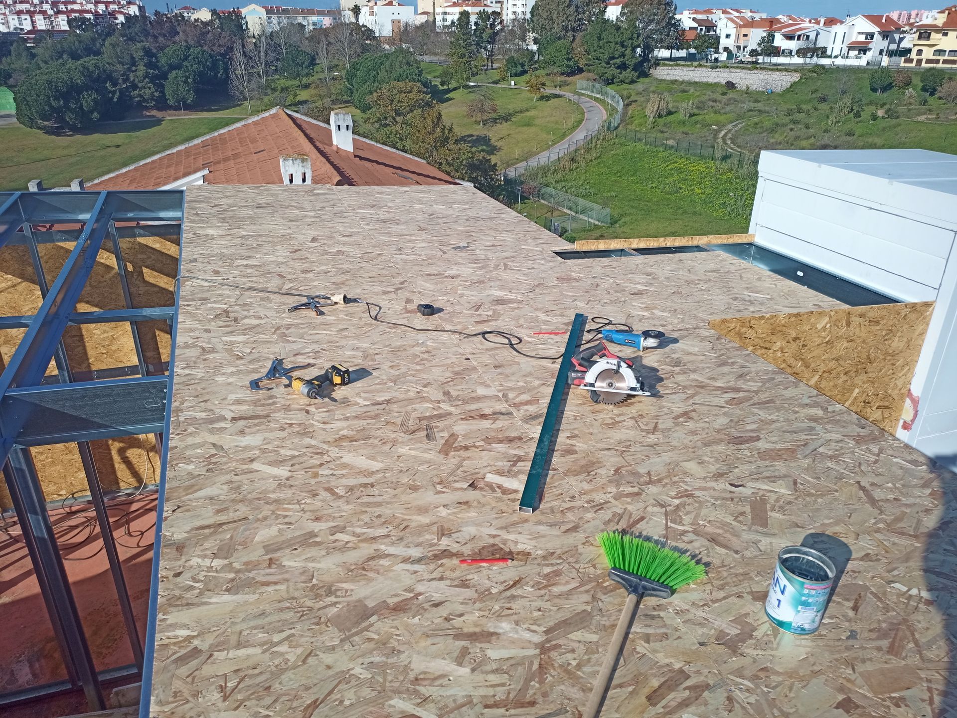 Canteiro de obras com painéis de OSB no telhado, estrutura metálica, ferramentas e vista de fundo com árvores e edifícios.