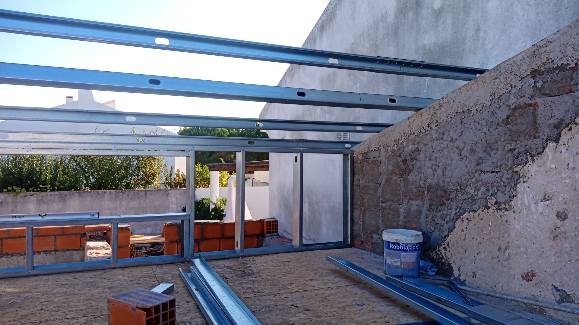 Canteiro de obras com estrutura metálica. Tijolos e materiais visíveis. Parede de concreto claro à direita.