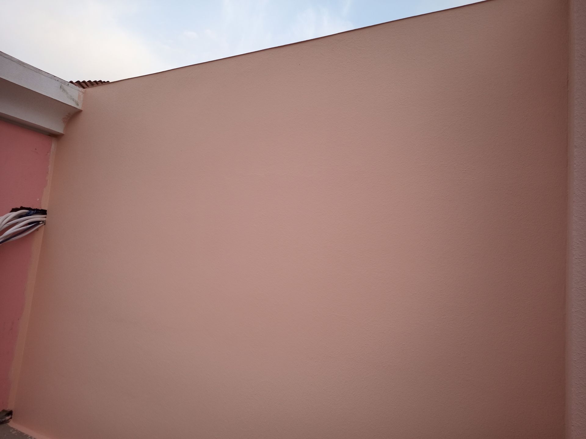 Parede rosa sob um céu azul claro com detalhes brancos na parte superior e fios expostos no lado esquerdo.