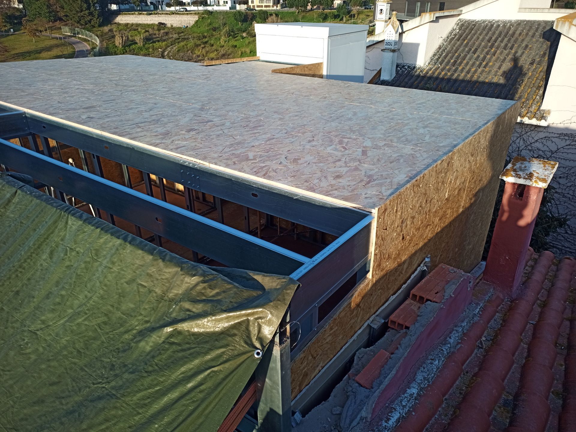Telhado plano em construção, parcialmente coberto por lona, ​​adjacente a um telhado de tijolos.