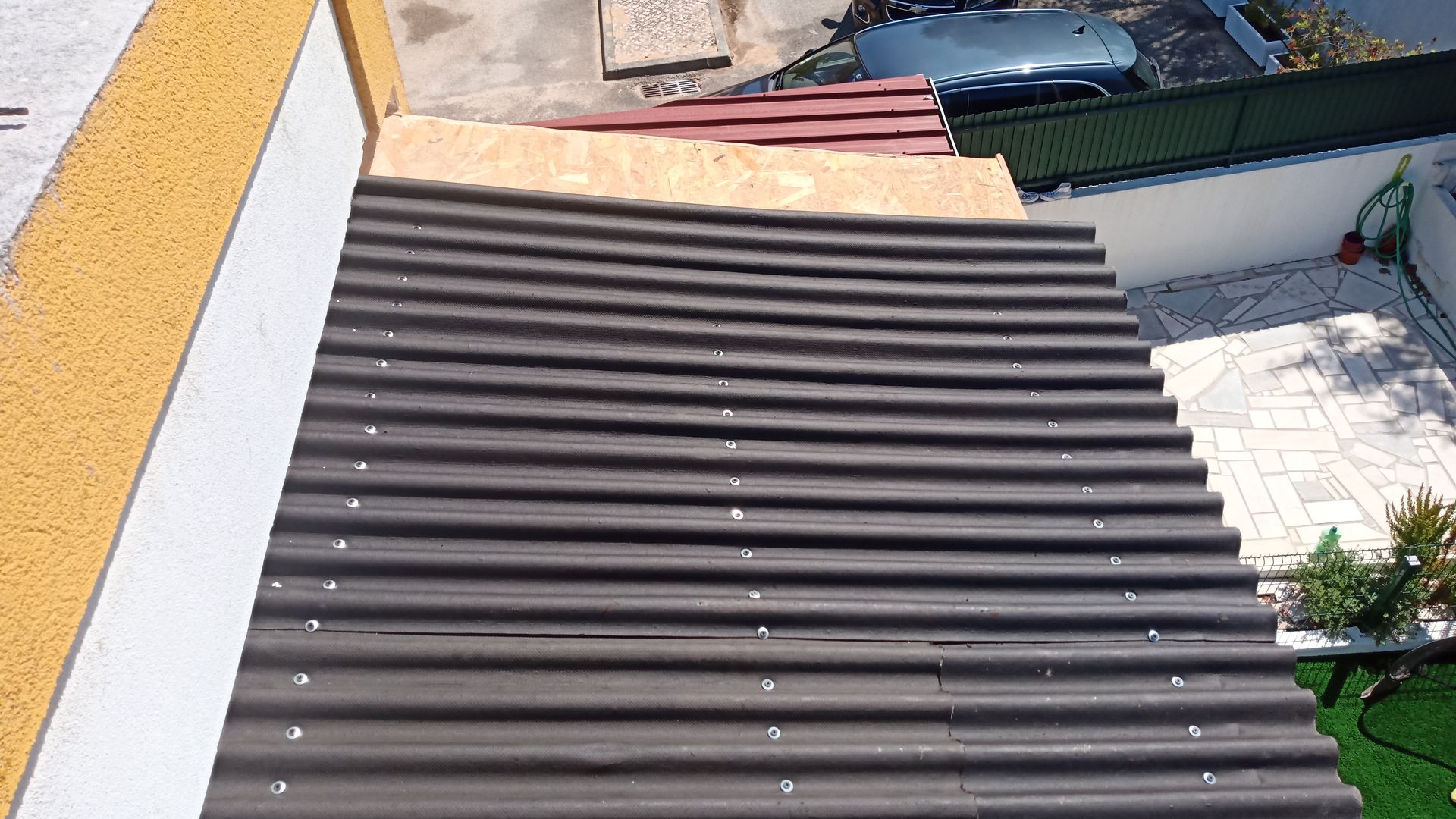 Vista aérea de um telhado escuro de chapa ondulada, uma parede bege e parte de um pátio branco com um carro ao fundo.