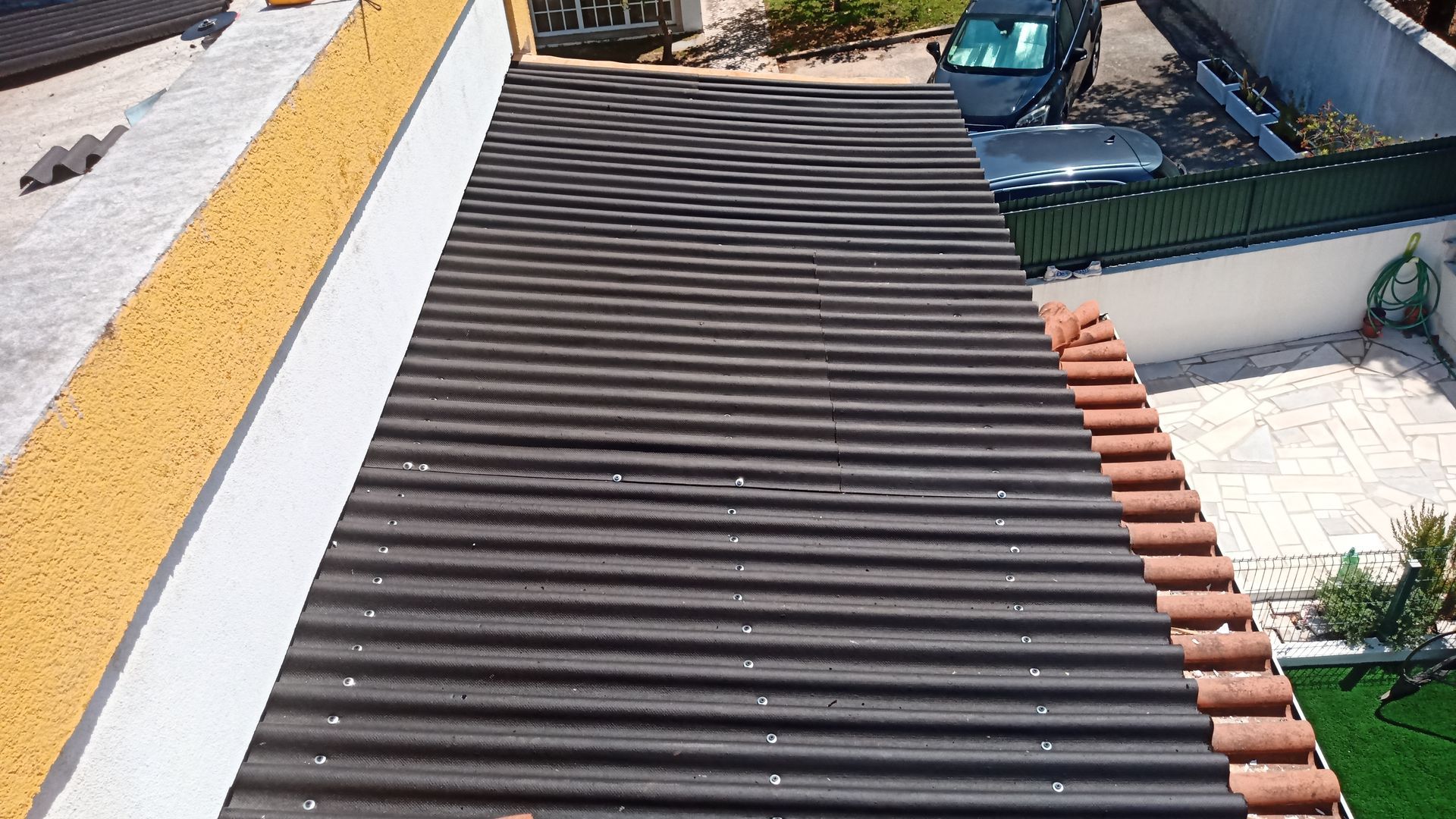 Vista de um telhado escuro de zinco ondulado, delimitado por uma parede amarela e telhas vermelhas. Uma cerca verde e um veículo são visíveis.