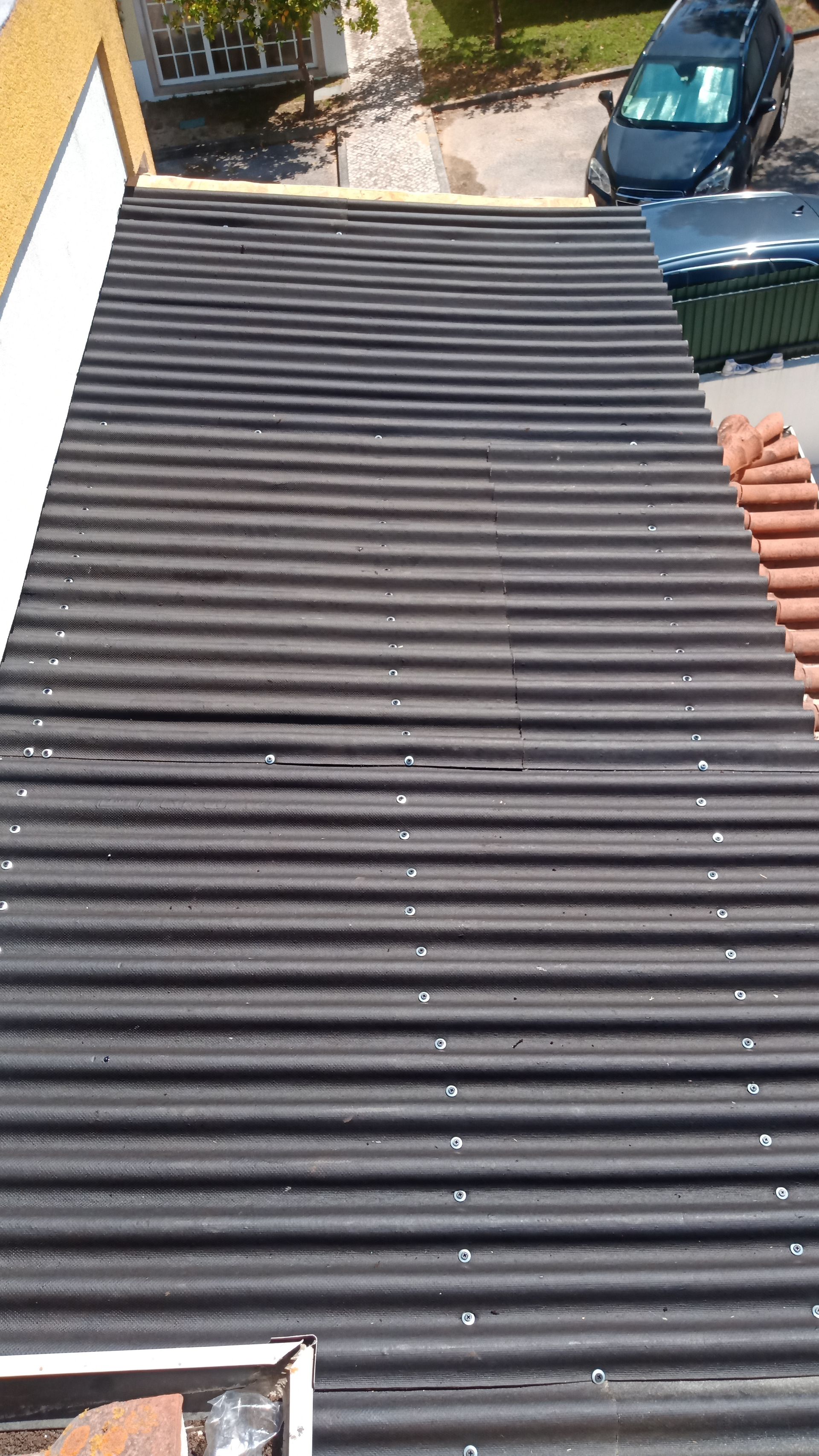 Vista de cima, uma cobertura preta ondulada. Um carro e uma lixeira são visíveis à direita.