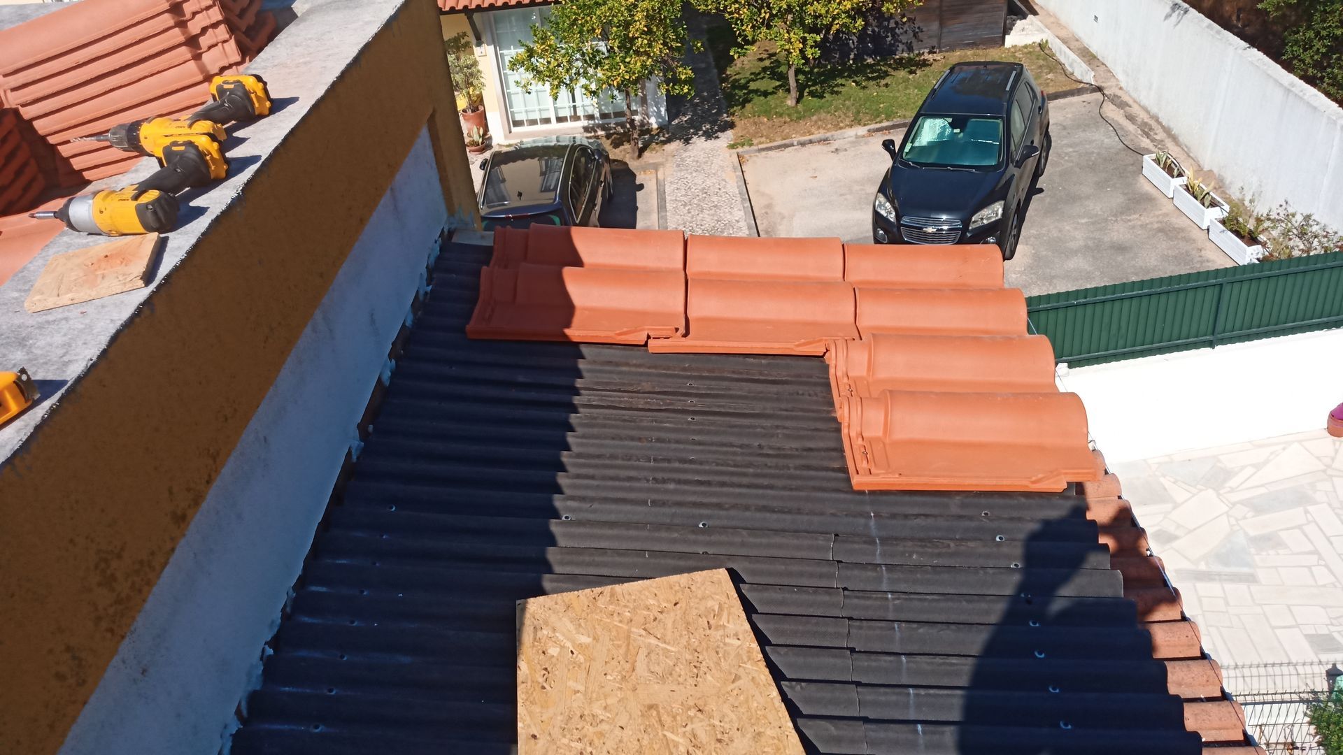Telhado com telhas laranja sendo instaladas. Subcobertura preta visível, ferramentas e carro ao fundo.