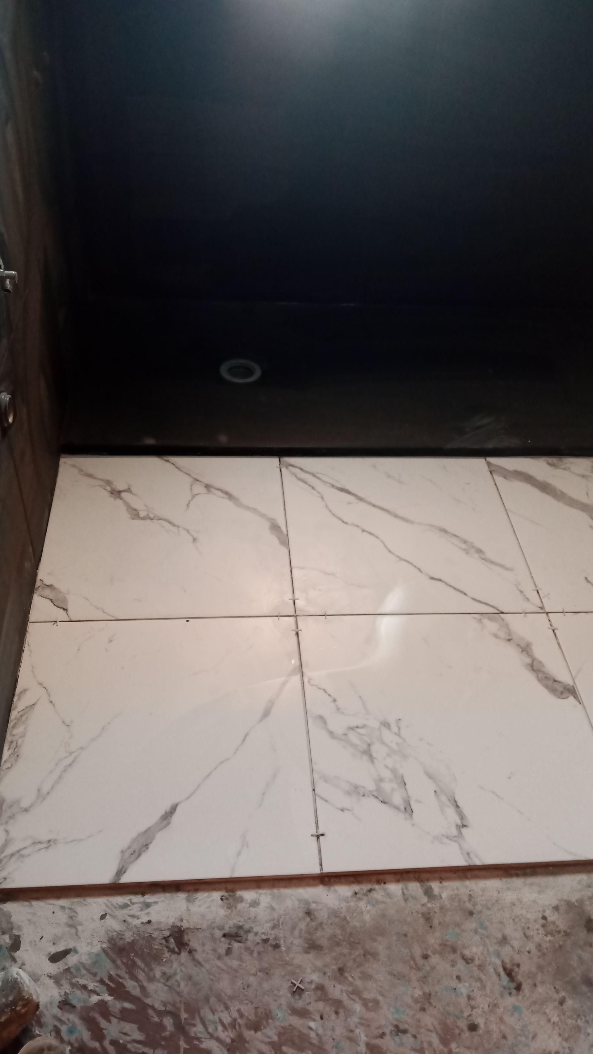Azulejos brancos com aparência de mármore no piso do banheiro, contra um teto escuro. Entulho de construção visível ao redor dos azulejos.