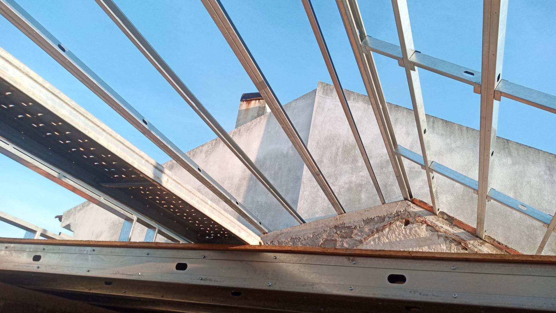 Estrutura metálica de uma construção contra um céu azul, obscurecendo parcialmente um edifício com chaminé.