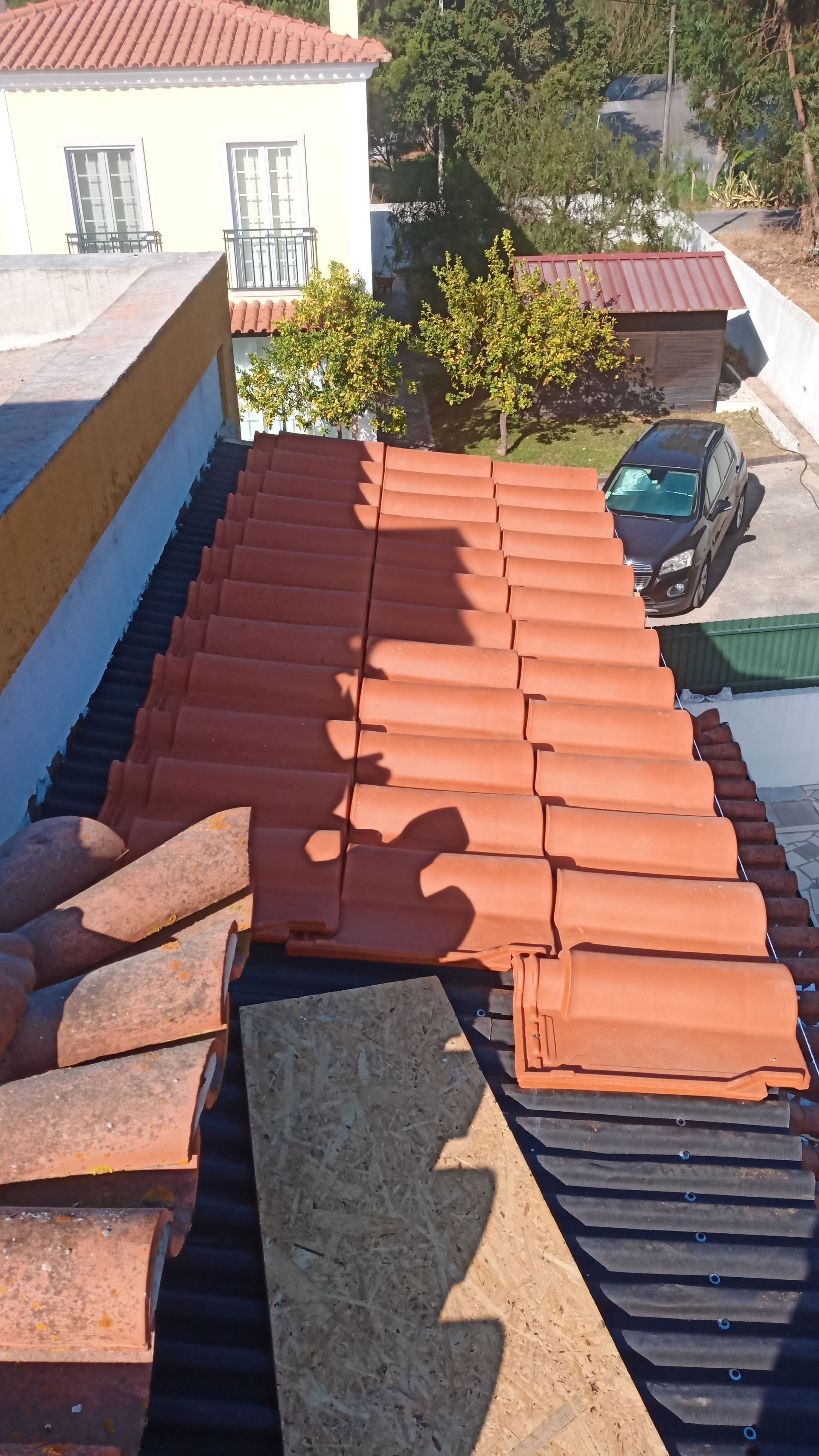 Telhado com telhas de barro laranja em fase de instalação. Uma casa e um carro são visíveis ao fundo.