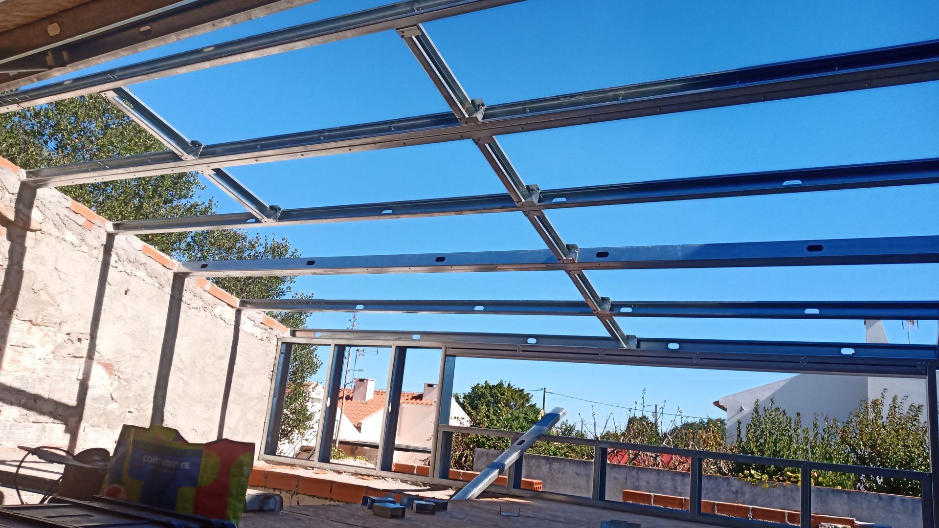 Estrutura metálica para um teto em construção contra um céu azul, com partes das paredes de tijolos visíveis.