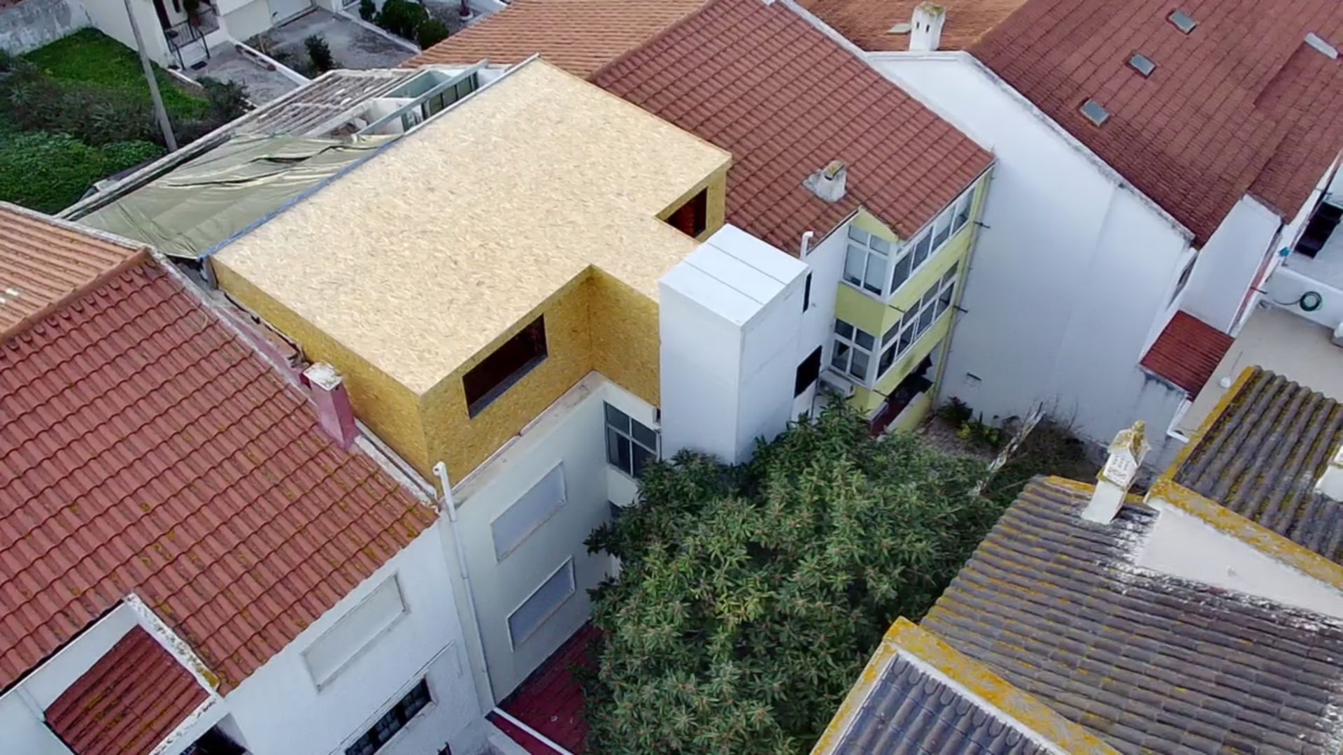 Vista aérea de edifícios com telhados de telha vermelha, uma estrutura de madeira clara e uma grande árvore verde.