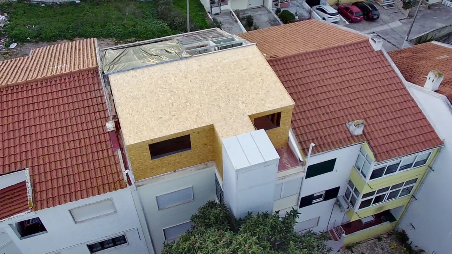 Vista aérea de um edifício em construção, com estrutura de madeira e casas circundantes com telhados de telha vermelha.
