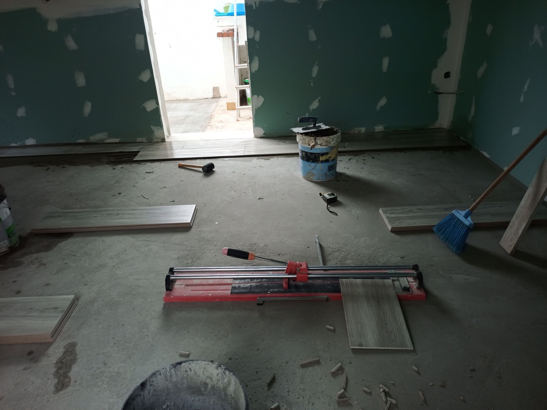 Sala em construção, com cortador de azulejos, azulejos e ferramentas sobre um piso de concreto. Paredes verde-azuladas e porta aberta.