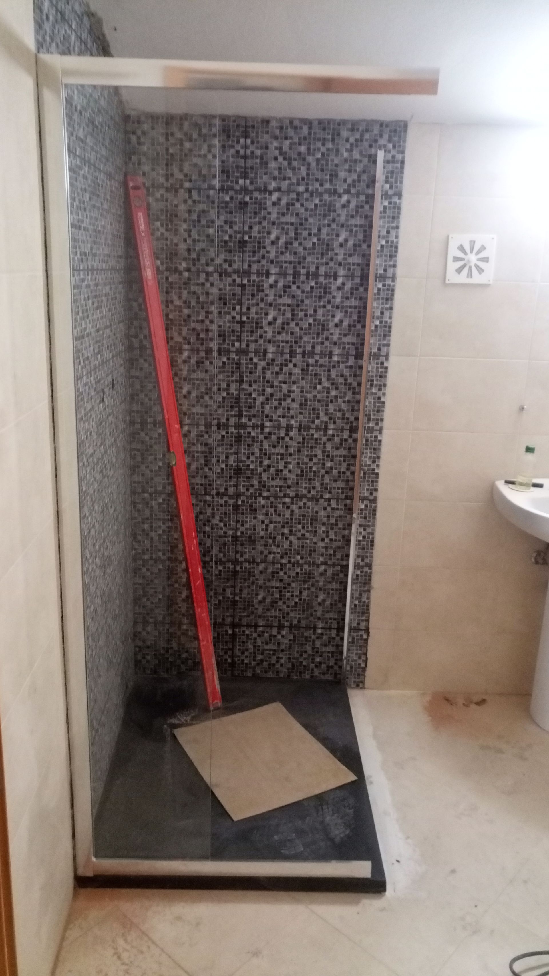 Box de chuveiro em construção, com azulejos pretos e cinzas, porta de vidro transparente e nível vermelho.