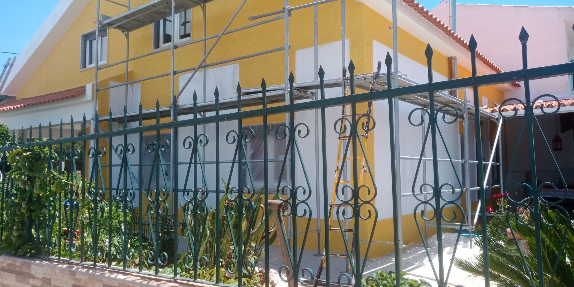 Casa amarela em reforma, parcialmente escondida por uma cerca de ferro verde-escura com detalhes ornamentais e folhagem exuberante.