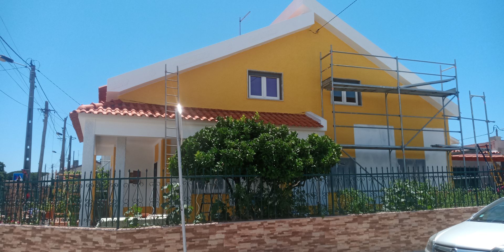 Casa amarela com andaimes, detalhes brancos e telhado de telhas vermelhas sob um céu azul e límpido.