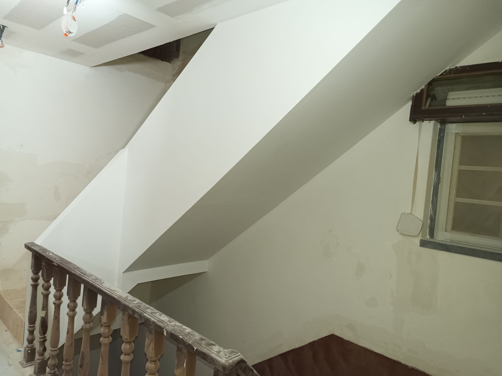 Escadaria com paredes brancas e corrimão de madeira. Janela com moldura escura à direita.