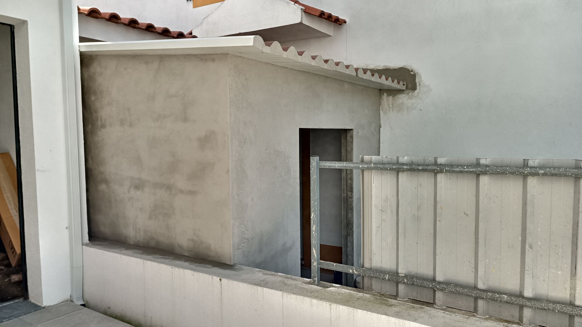 Estrutura de concreto cinza com uma porta, ao lado de uma parede branca e uma cerca baixa.