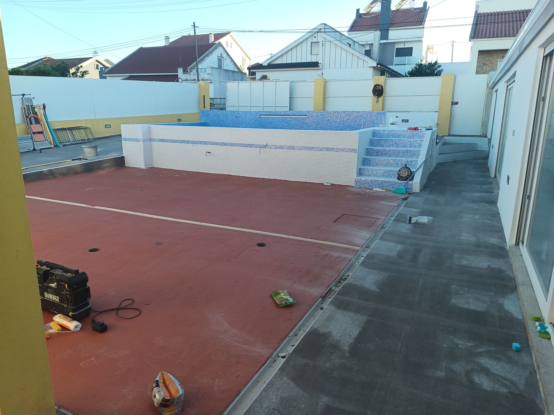 Espaço exterior com superfície vermelha, uma piscina em construção e edifícios ao fundo.