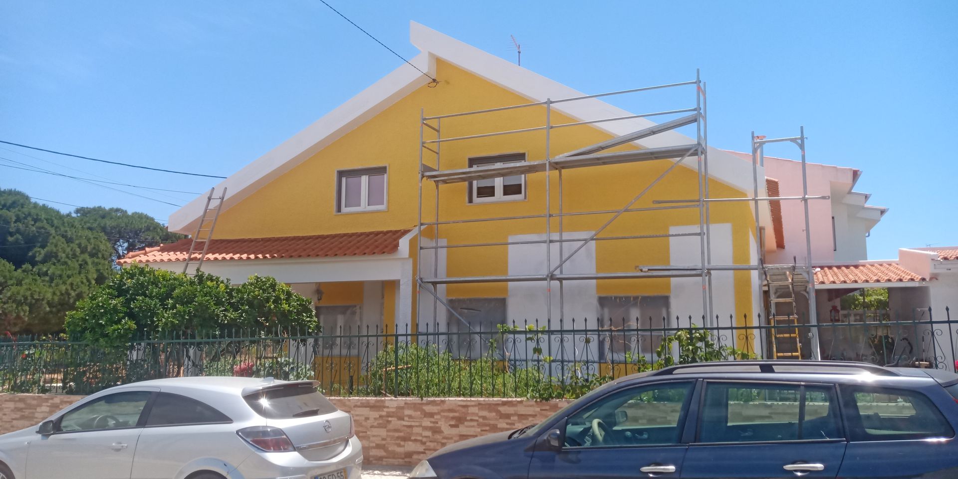 Casa amarela com andaimes, carros estacionados na rua. Dia ensolarado.