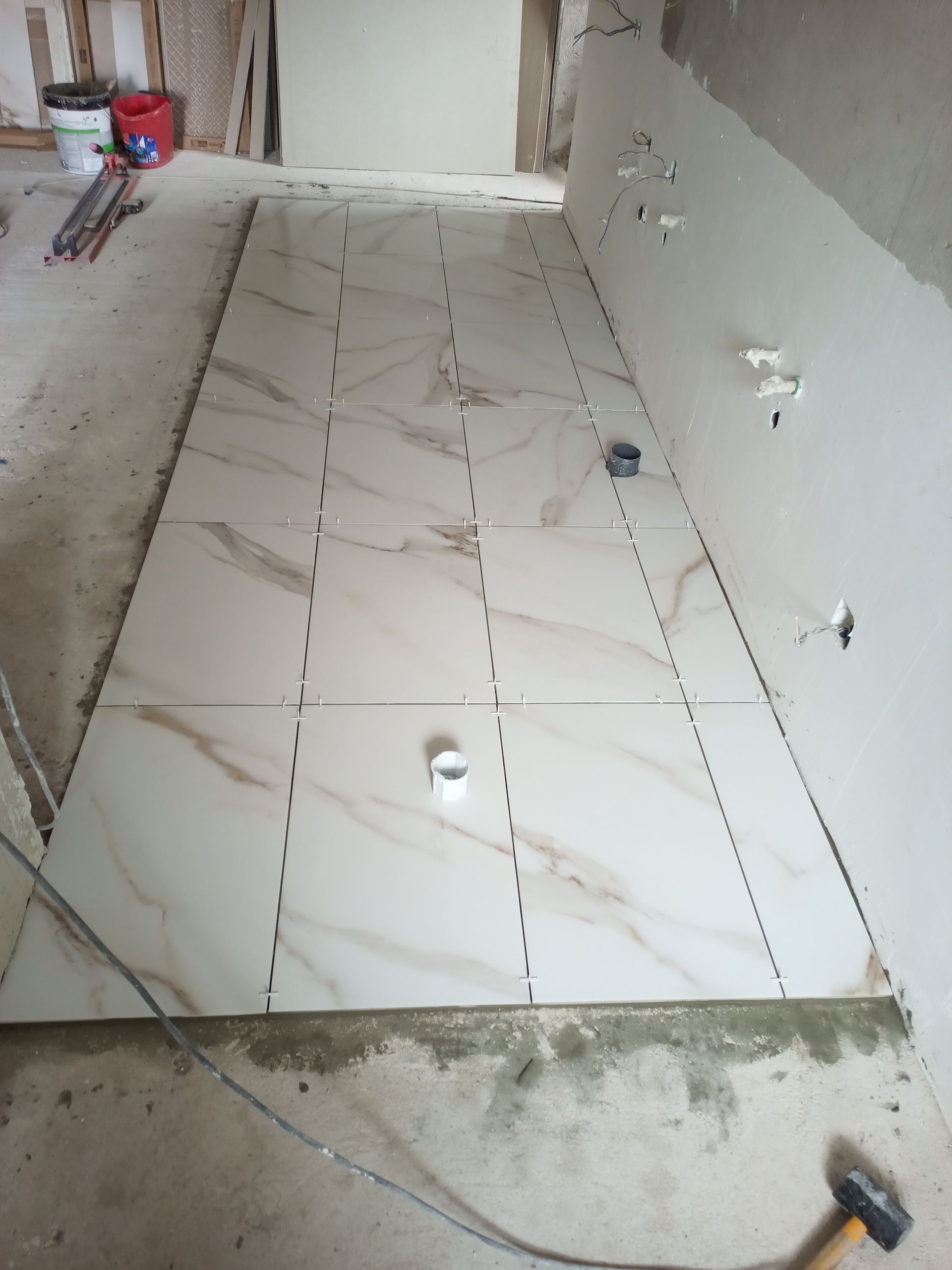 Colocação de azulejos no piso em andamento; grandes azulejos retangulares dispostos com espaçadores. Padrão marmorizado em branco e bege.