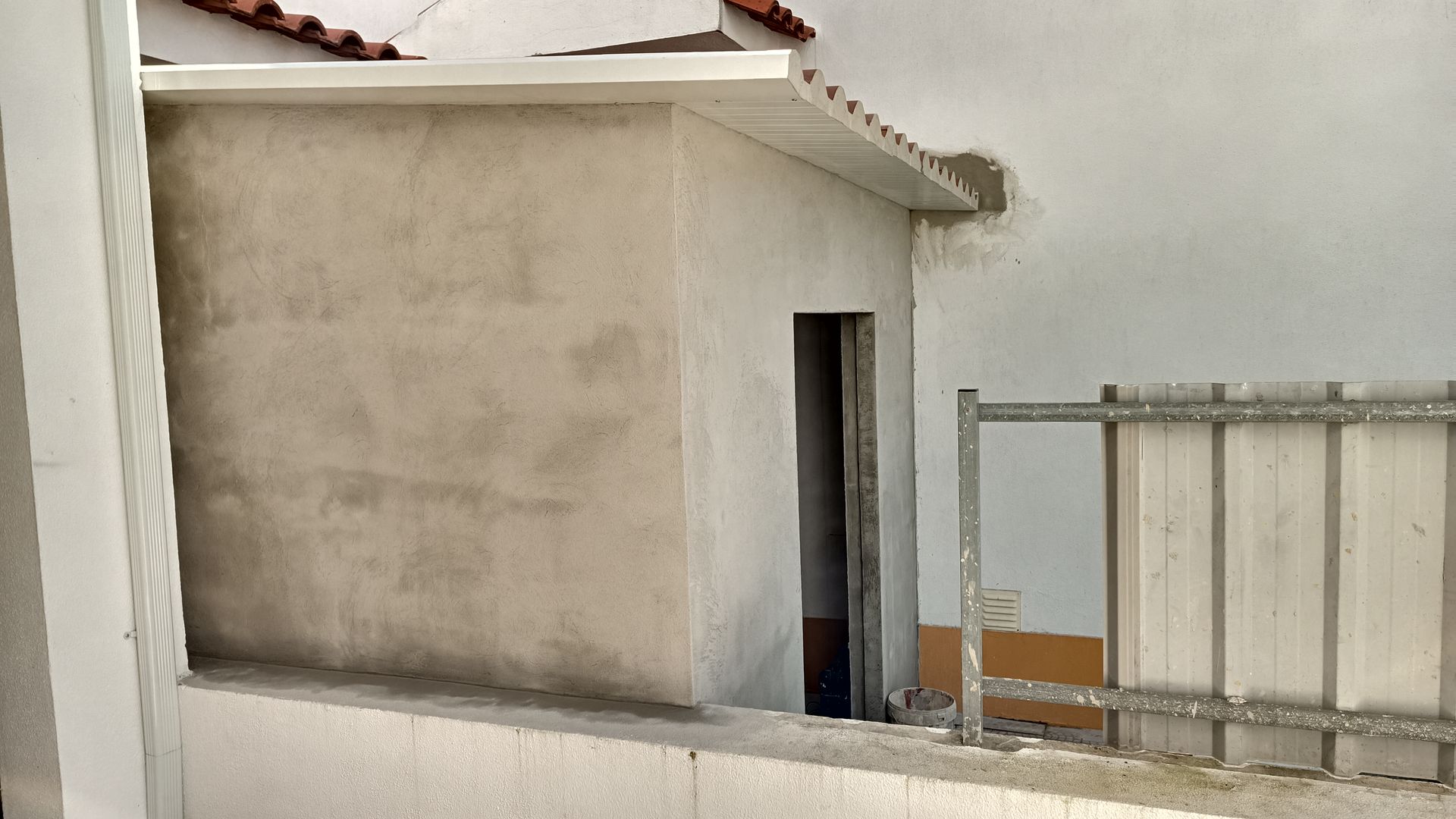 Estrutura de concreto com porta aberta, detalhes em branco e vista parcial do telhado de telhas.