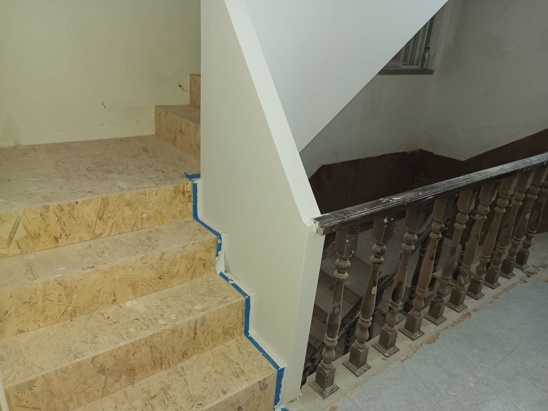 Escadaria com degraus de mármore, corrimão de madeira e paredes bege. Fitas azuis marcam os locais onde a pintura está sendo feita.