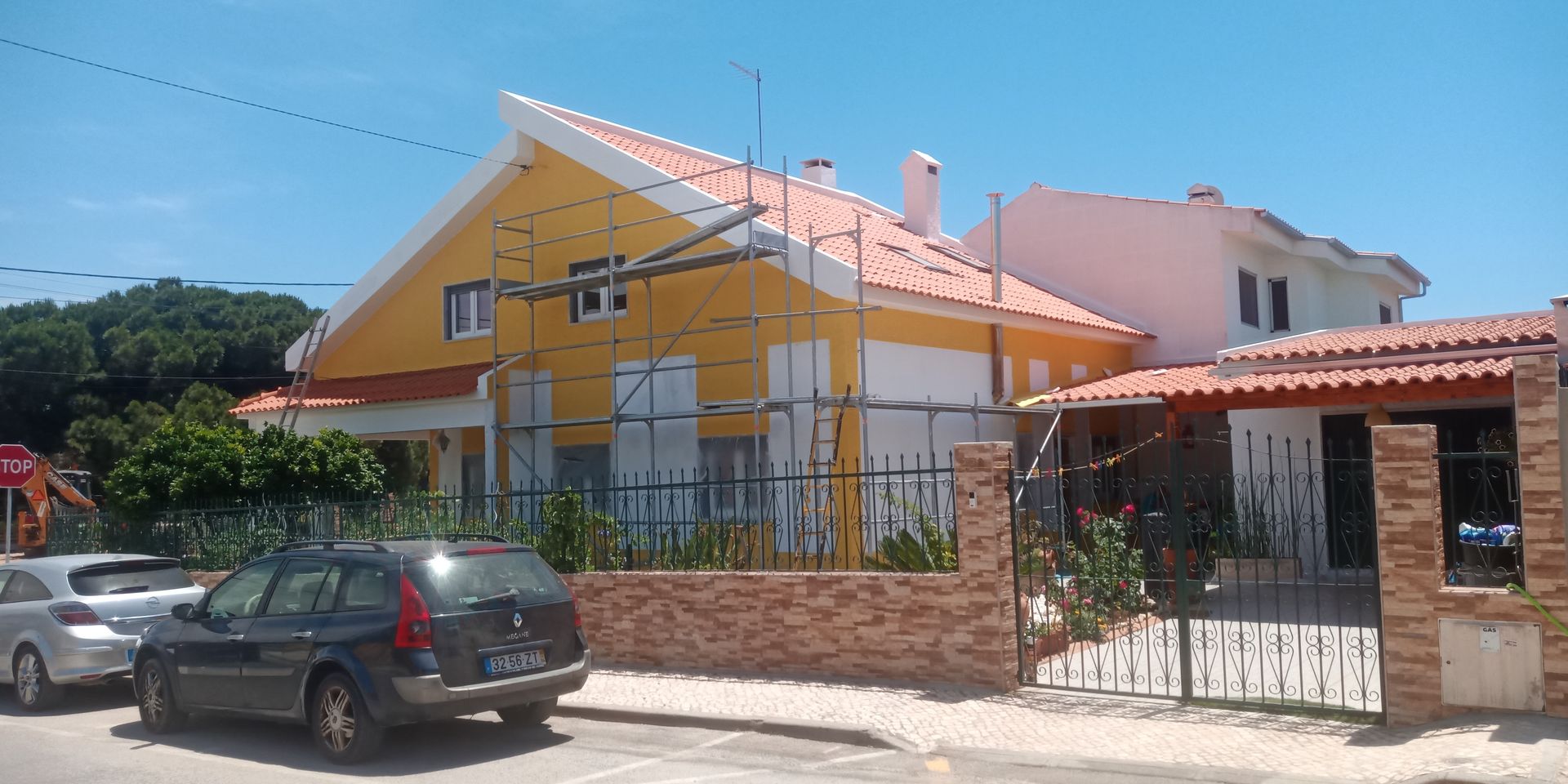 Casa amarela com telhado vermelho e andaimes; carros estacionados na rua.