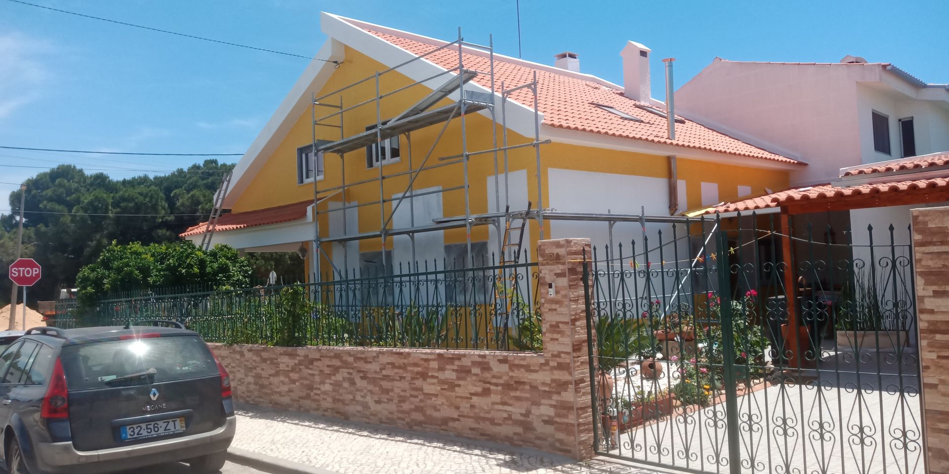 Casa amarela com telhado laranja em construção, cercada por muro e vegetação, com um carro estacionado em frente.