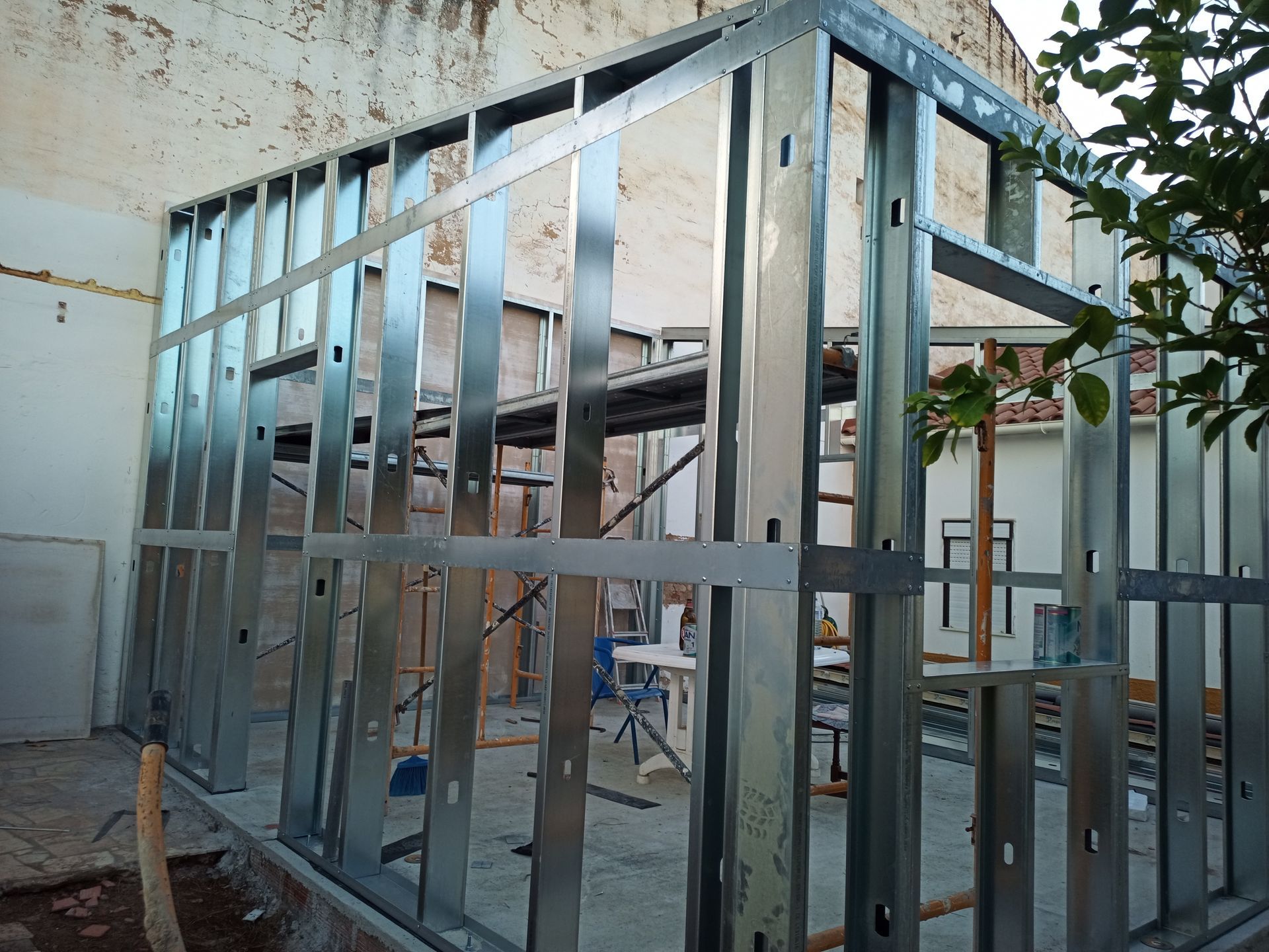 Estrutura metálica de uma construção em andamento ao ar livre, com um prédio branco ao fundo.