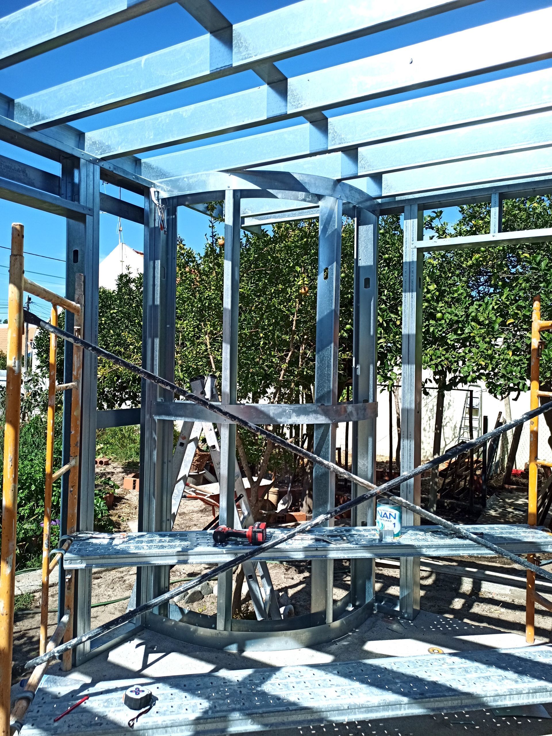 Estrutura de aço de um edifício em construção, com vista para árvores ao fundo, em ambiente externo.